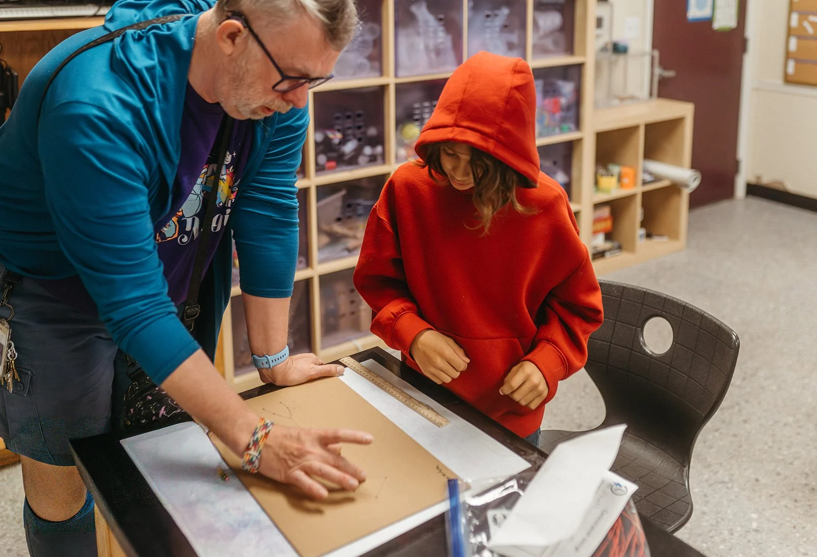 An older man with glasses and a beard demonstrates something on a wooden board to a young girl in a red hoodie, in a room with wooden shelves filled with small containers and items.