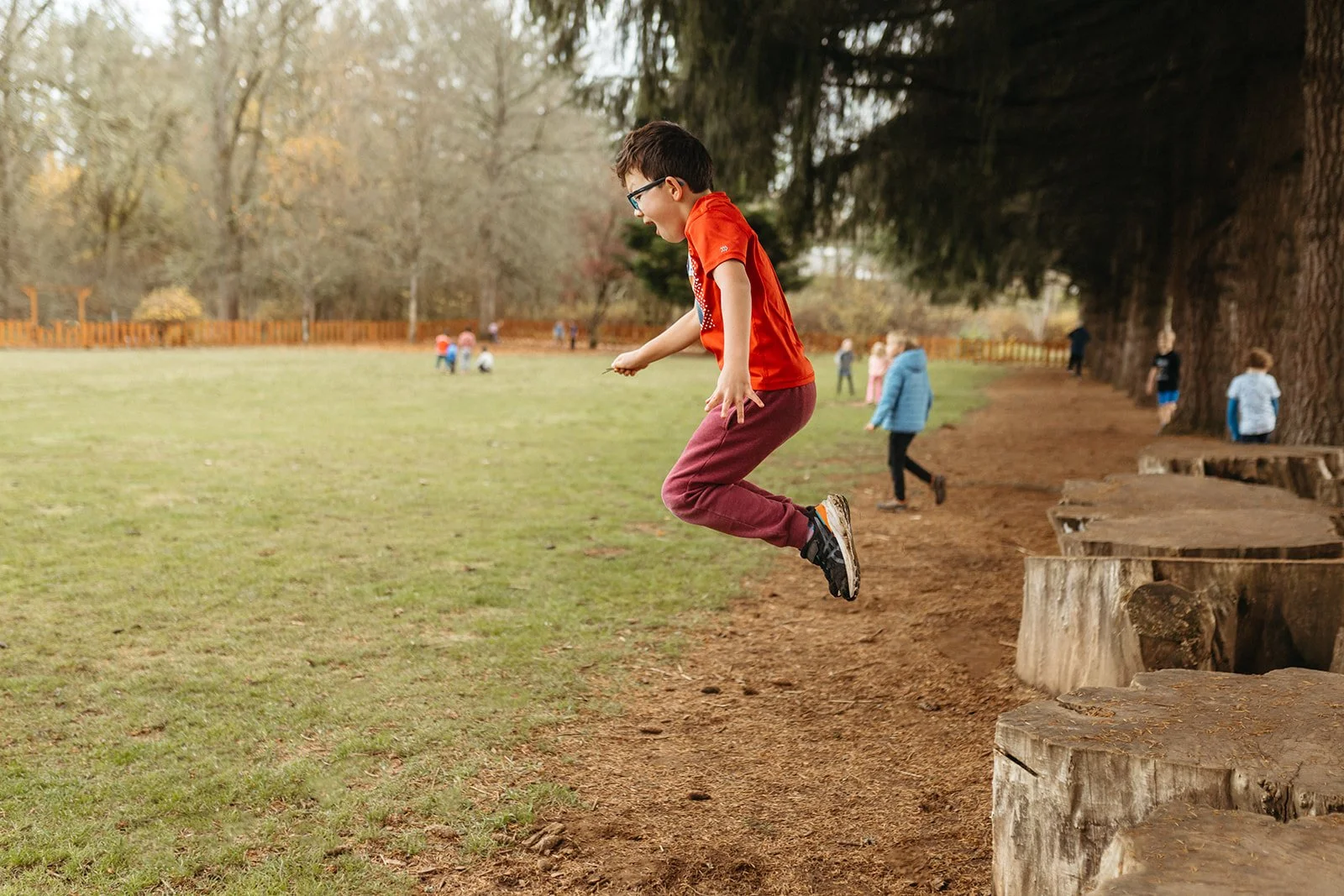 A boy in a red shirt and maroon pants jumping in the air, in a park with others and large trees in the background.