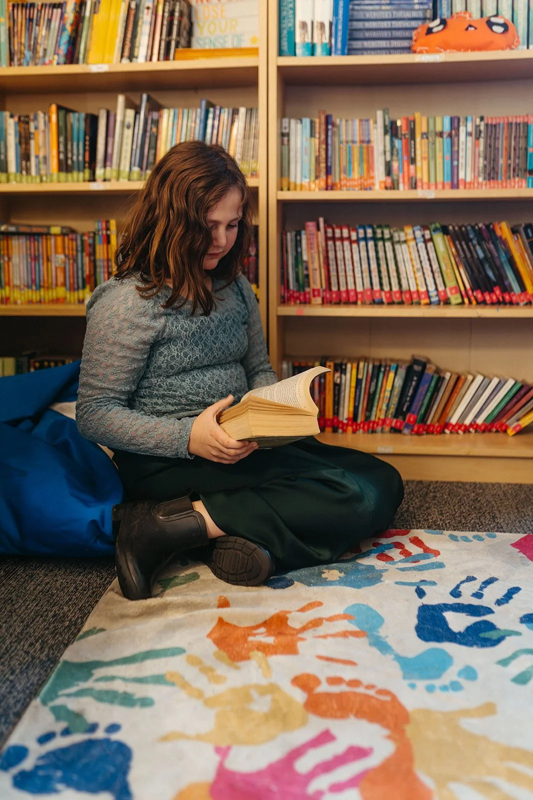 A young girl with wavy brown hair sitting on the carpet in a library, reading a book, with bookshelves filled with colorful books behind her.
