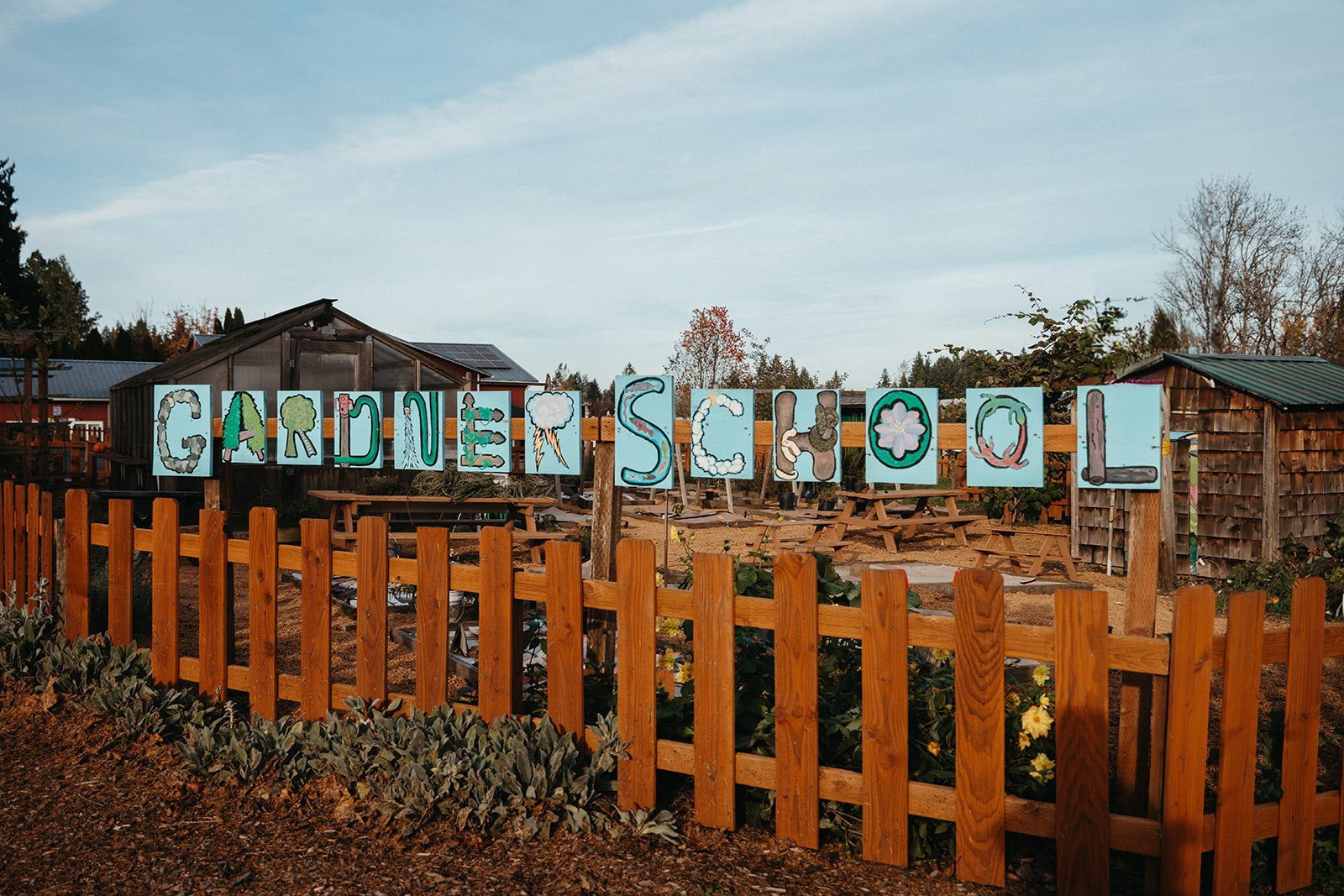 Sign spelling 'GARDEN SCHOOL' with letters decorated as nature and weather elements, inside a garden area with wooden fences, picnic tables, and small trees.