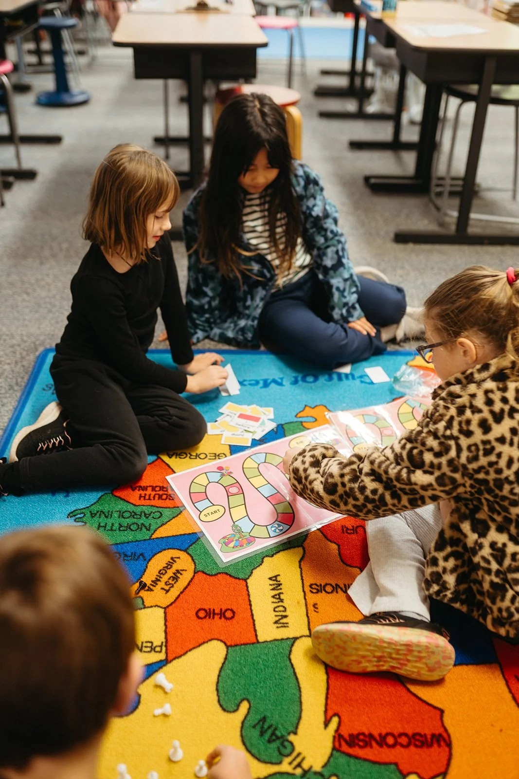 Children playing a board game on a colorful map-themed rug in a classroom.
