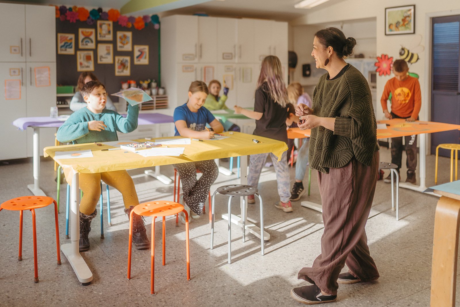 Children and a woman in a classroom with colorful tables and chairs, engaging in an arts and crafts activity.