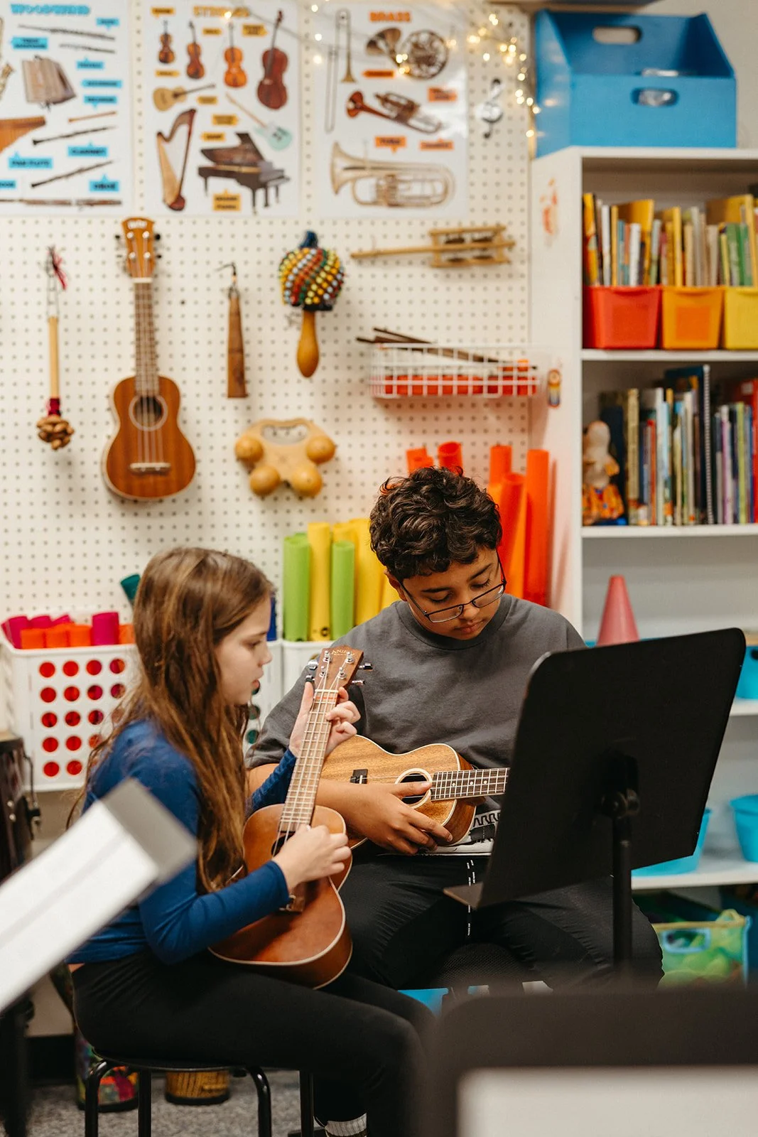 Two children sitting in a music classroom playing ukuleles, with musical instruments and books on the wall behind them.