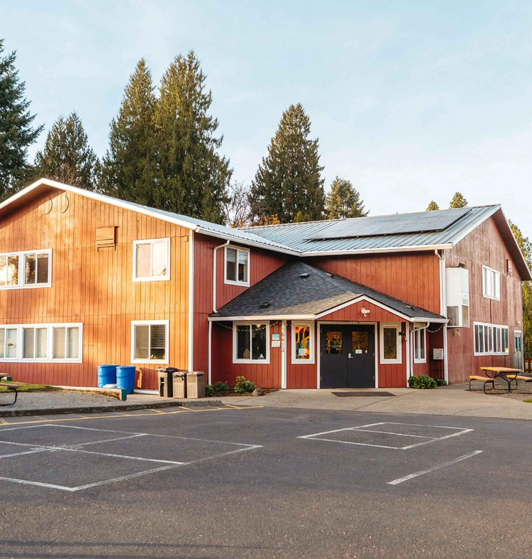 A wooden house with multiple windows and a gray metal roof, surrounded by tall pine trees, with a parking lot in front that has empty parking spaces and trash bins.