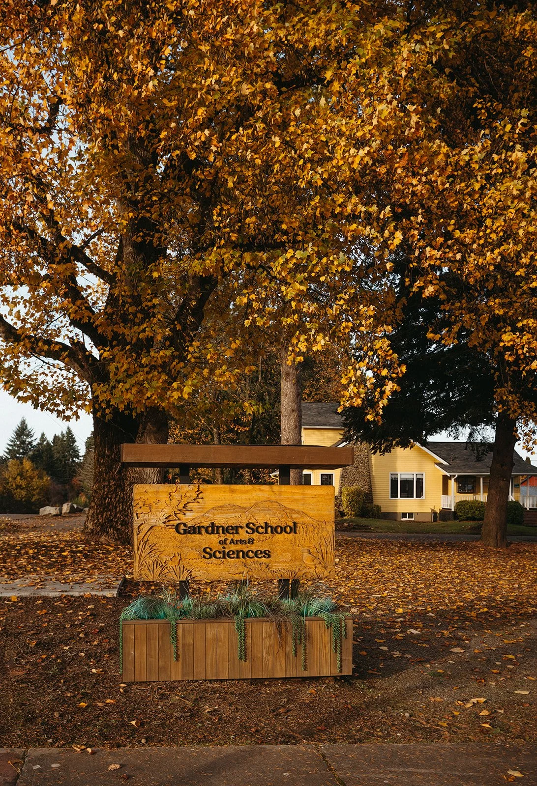 A wooden sign that reads 'Gardner School of Arts & Sciences' is located in front of a large tree with yellow and orange autumn leaves. In the background, there is a yellow house with white trim and a small porch.