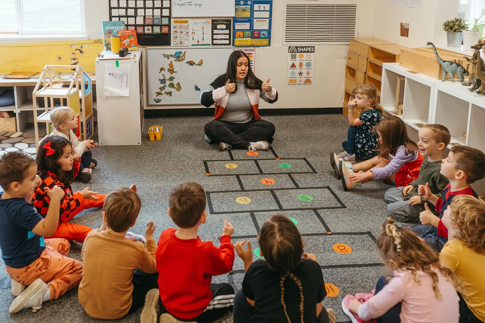 A teacher sits on the floor in a circle with young children in a classroom. The children are sitting on the carpet, facing the teacher, who is using a hopscotch game laid out on the floor for instruction or activity. The classroom has educational posters, toys, and decorations.