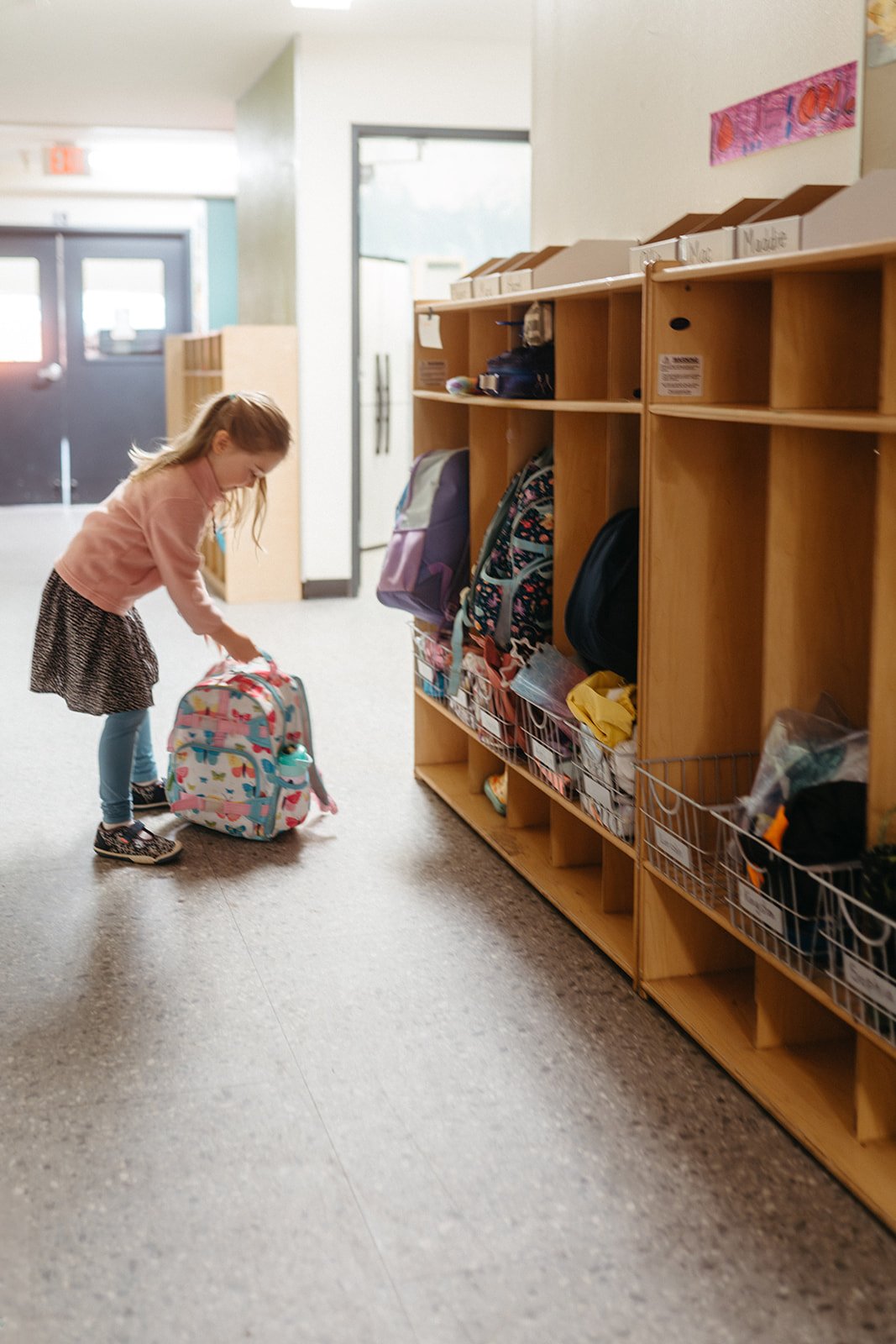 A young girl is bending over to pick up a colorful backpack in a school hallway with cubbies and backpacks on the shelves.