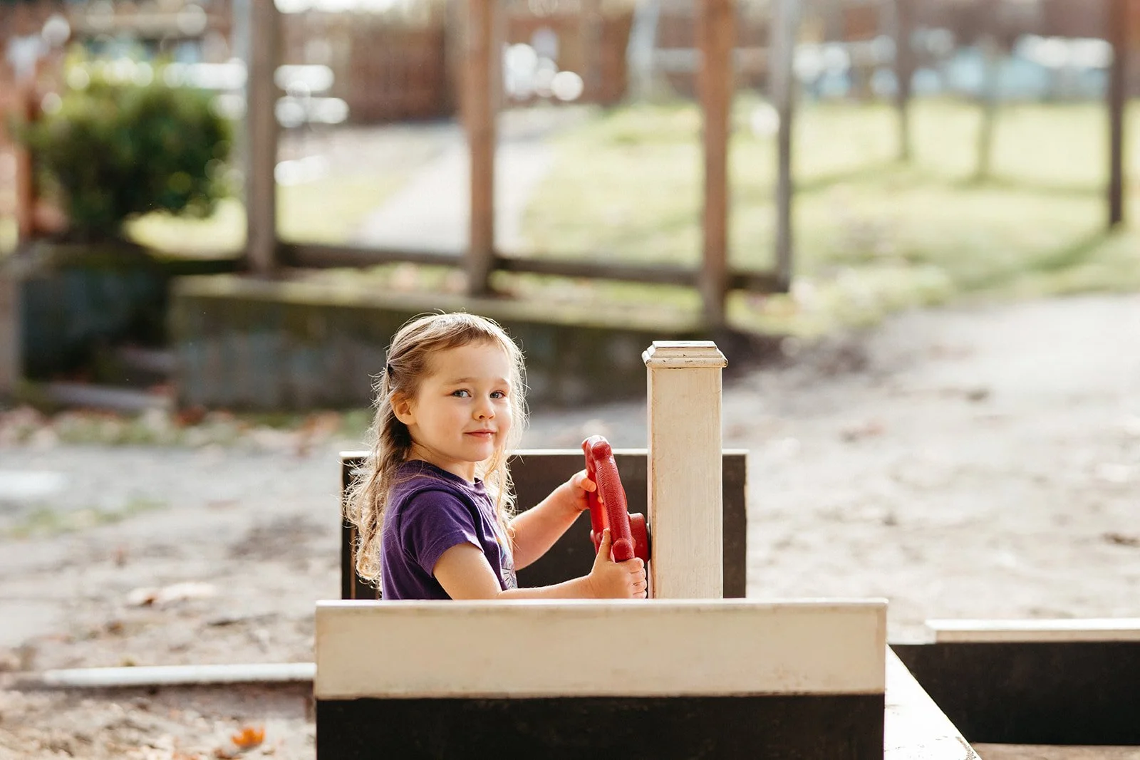 A young girl with long, curly blonde hair and a purple shirt sitting on a small wooden boat or ride, holding onto the steering wheel, outdoors with blurry trees and a wooden fence in the background.
