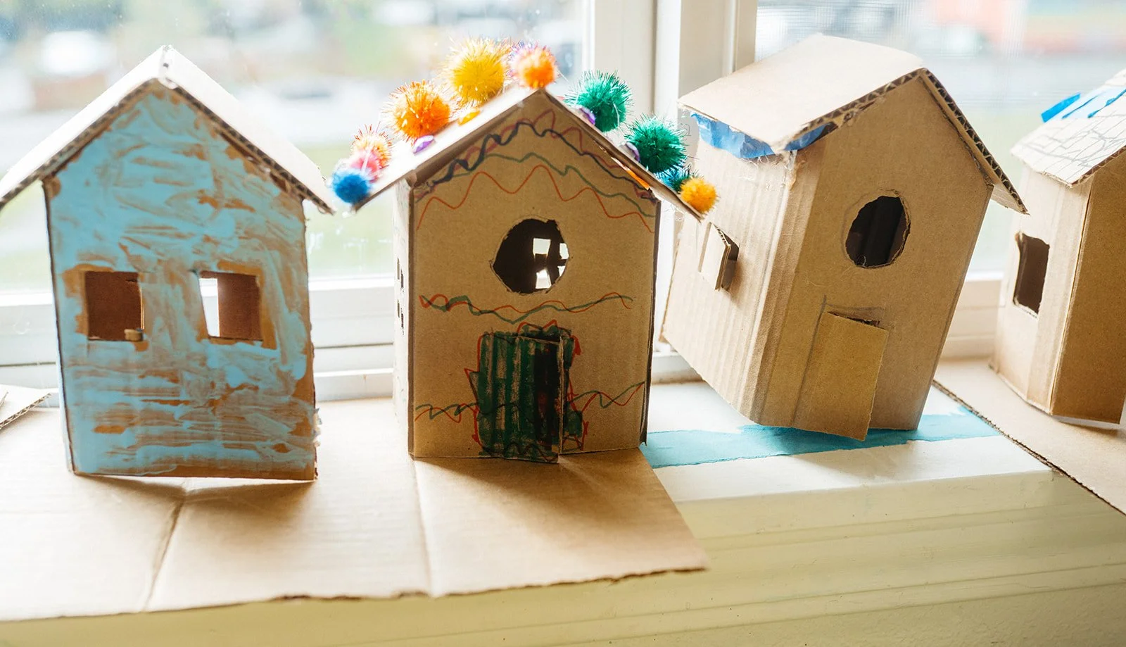 Cardboard houses decorated with paint, colorful pom-poms, and crayon drawings, arranged on a windowsill.