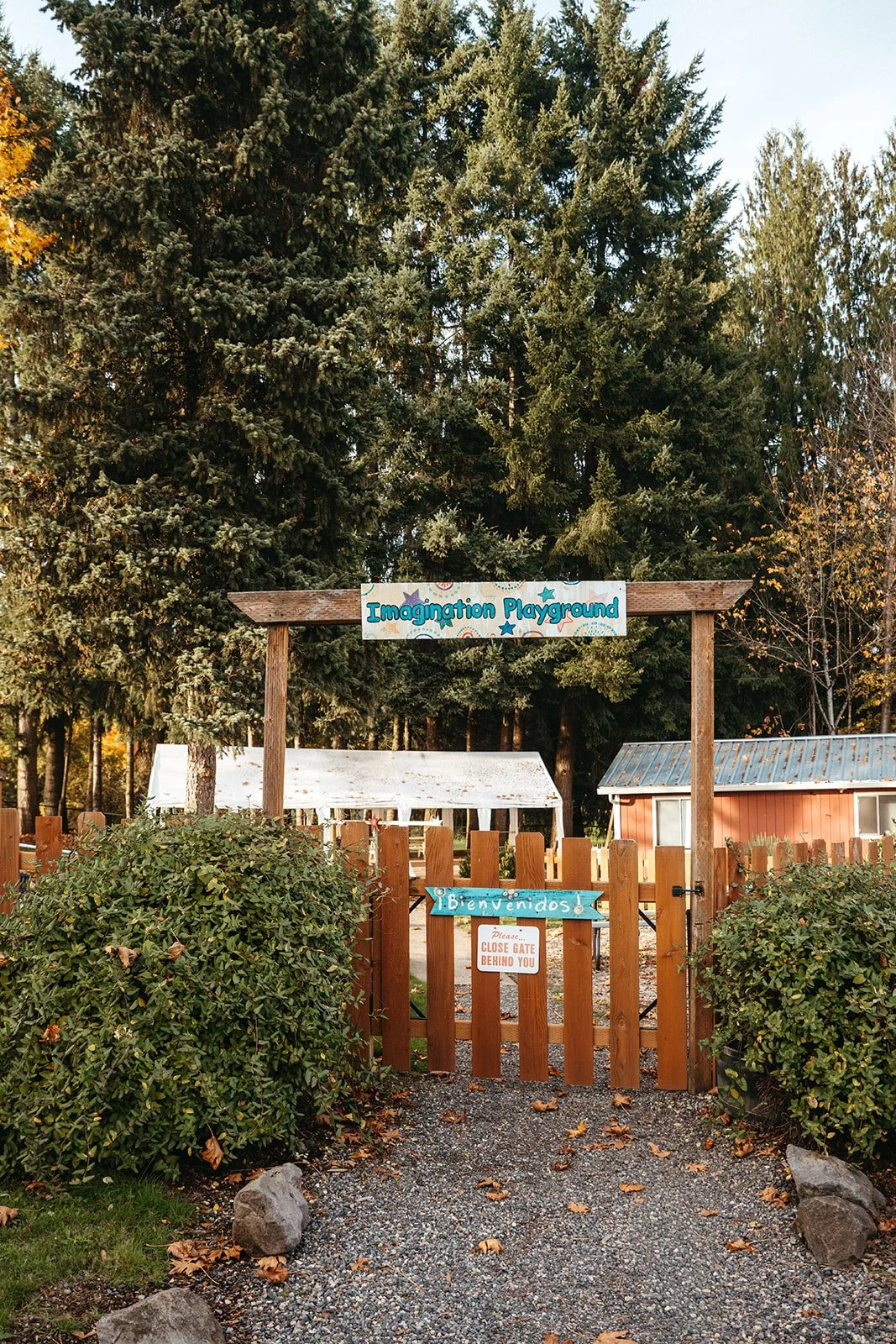 Entrance to Imagination Playground with a wooden gate, signs in English and Spanish, surrounded by greenery and trees.