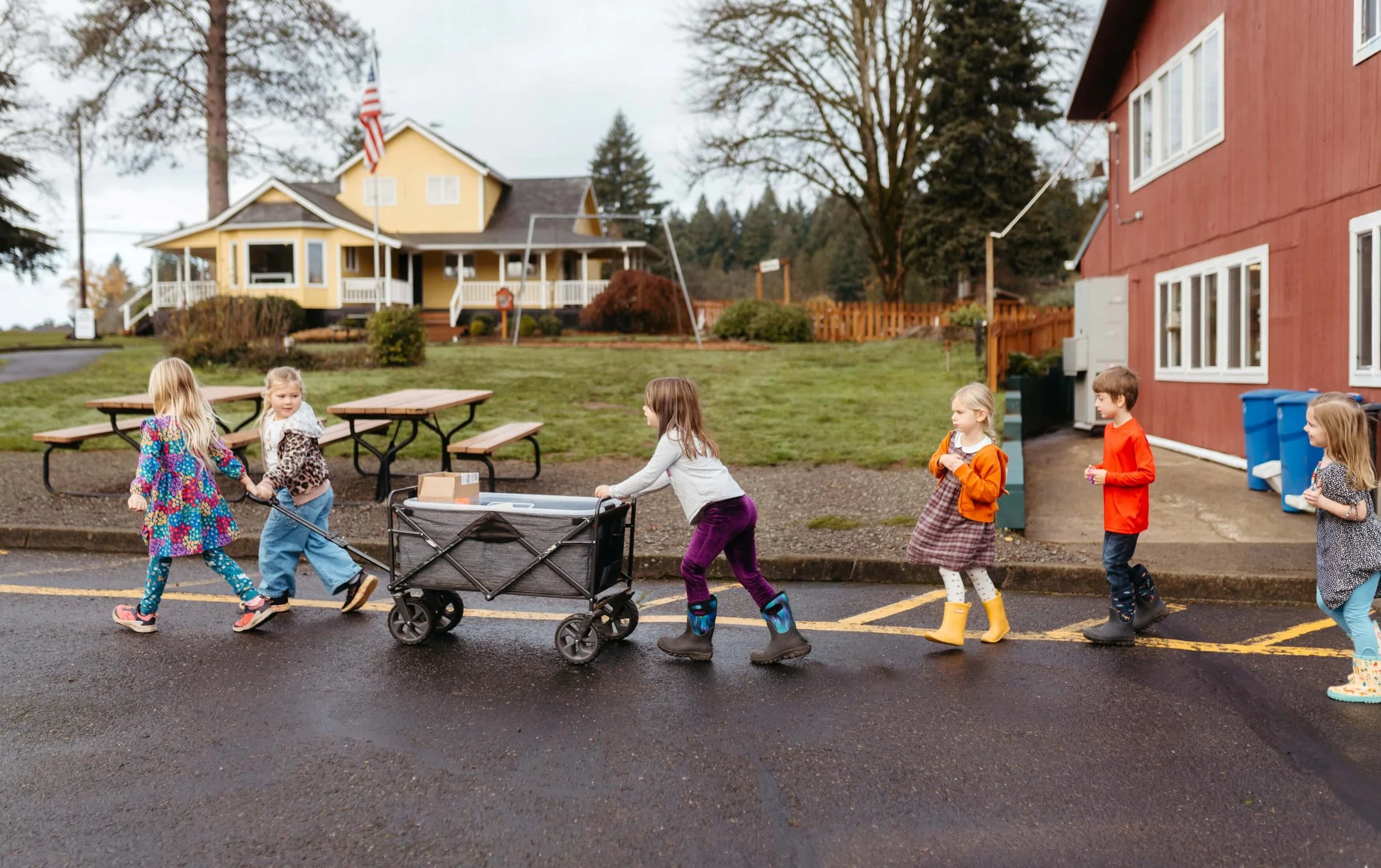 Children walking on a street in front of a red building, with a yellow house and trees in the background.