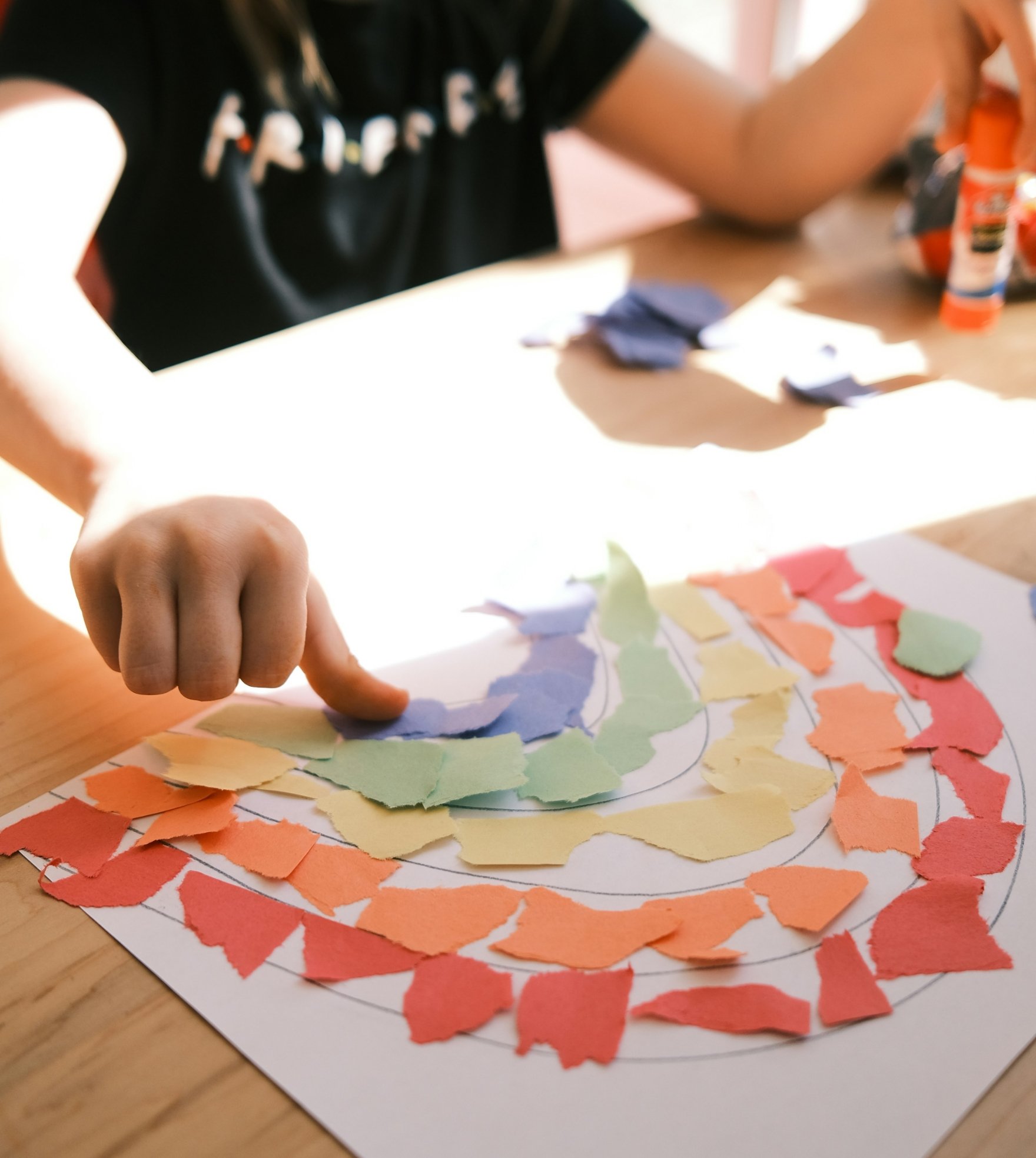 A child's hand pointing at a colorful paper collage on a piece of paper, with torn paper pieces in orange, yellow, green, and purple arranged in a pattern around the paper.