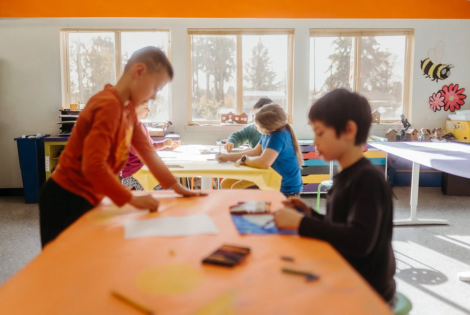 Children working on arts and crafts projects in a classroom with large windows, decorations, and sunlight streaming in.