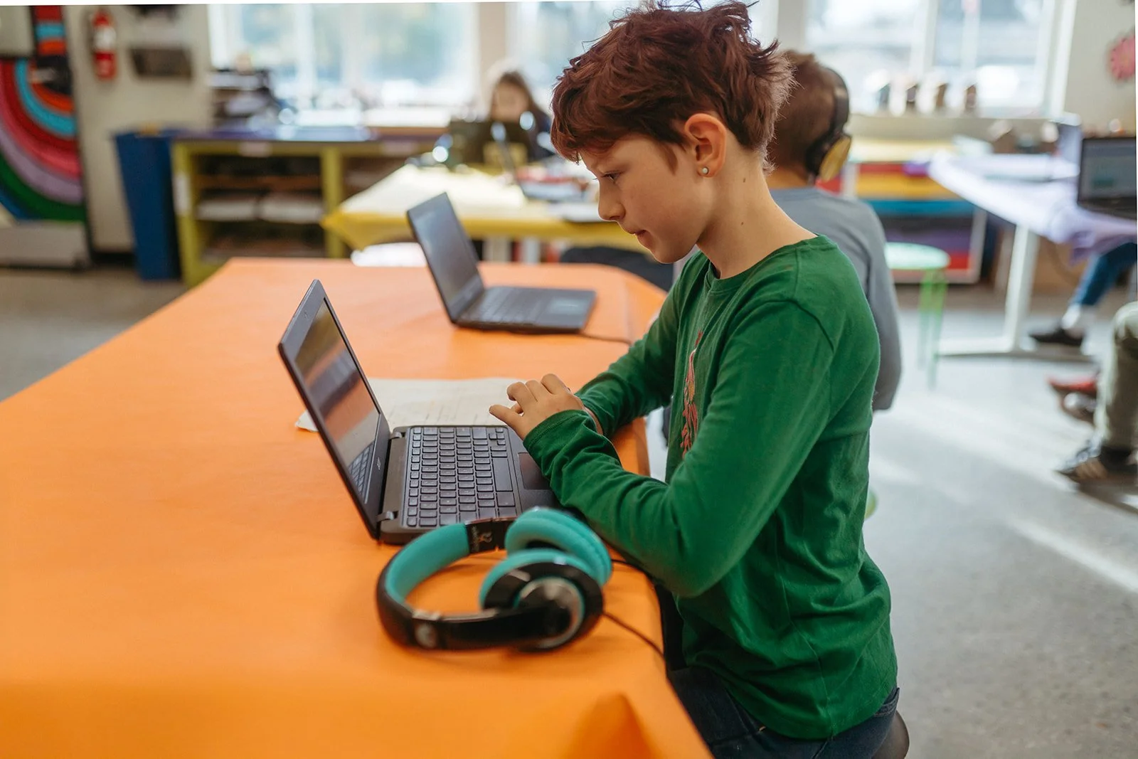 A girl with short reddish hair sitting at a school desk using a laptop, with headphones nearby, in a classroom setting with other students and colorful classroom decorations in the background.