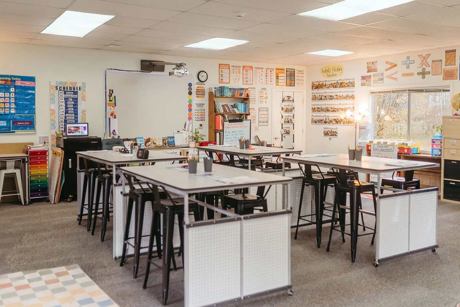Empty classroom with tables and chairs, educational posters on the walls, and sunlight coming through windows.
