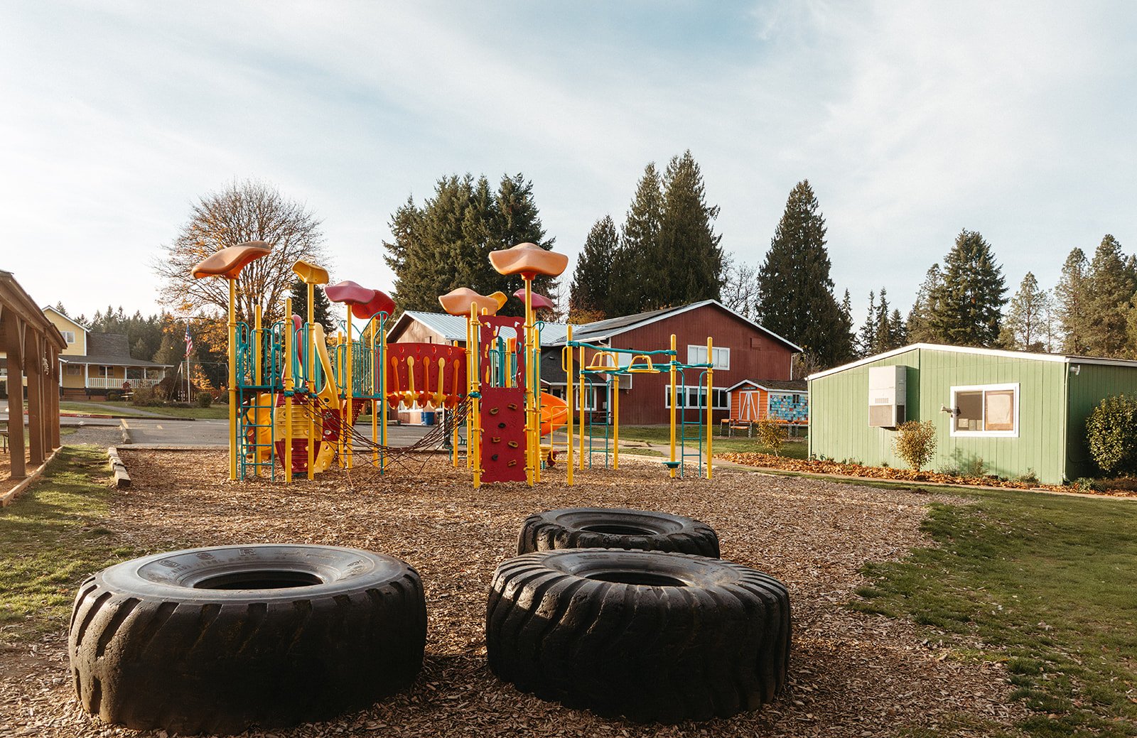 Colorful playground with slides, climbing structures, and tires in a park, surrounded by trees and small buildings.