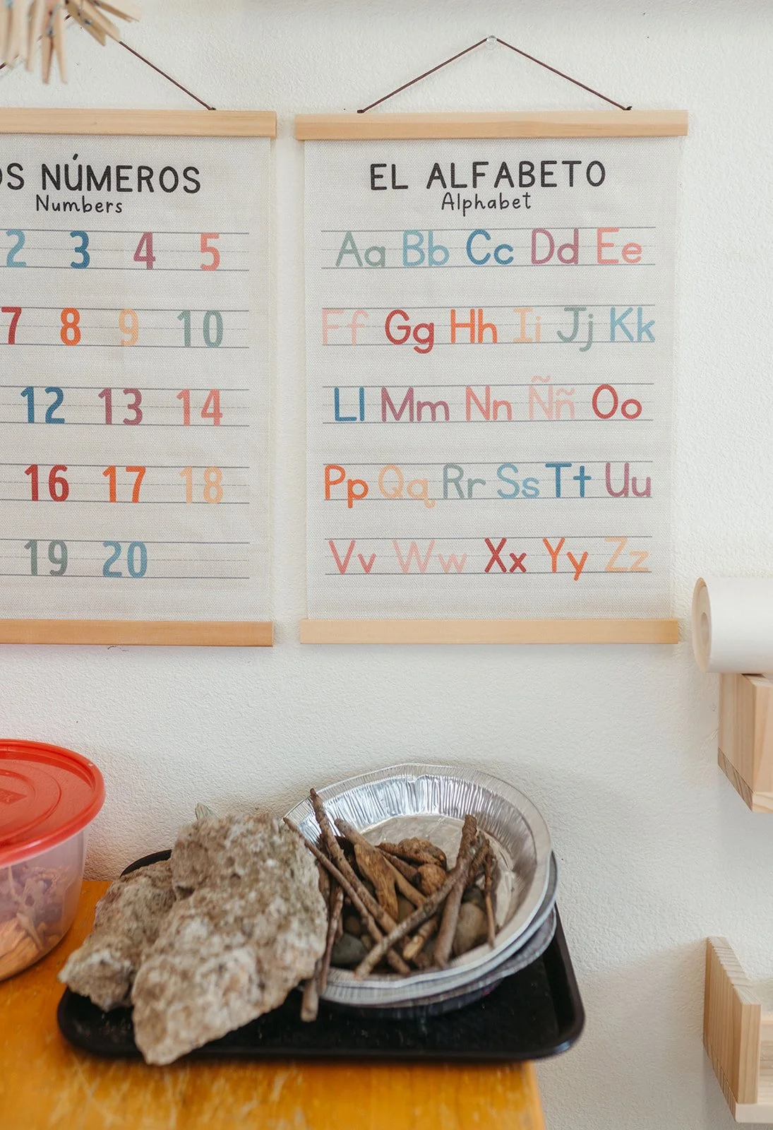 A wall with two educational posters, one showing numbers 1 to 20 and the other showing the alphabet. Below the posters, there is a black tray with rocks and small sticks on a yellow table, with a red container partly visible.