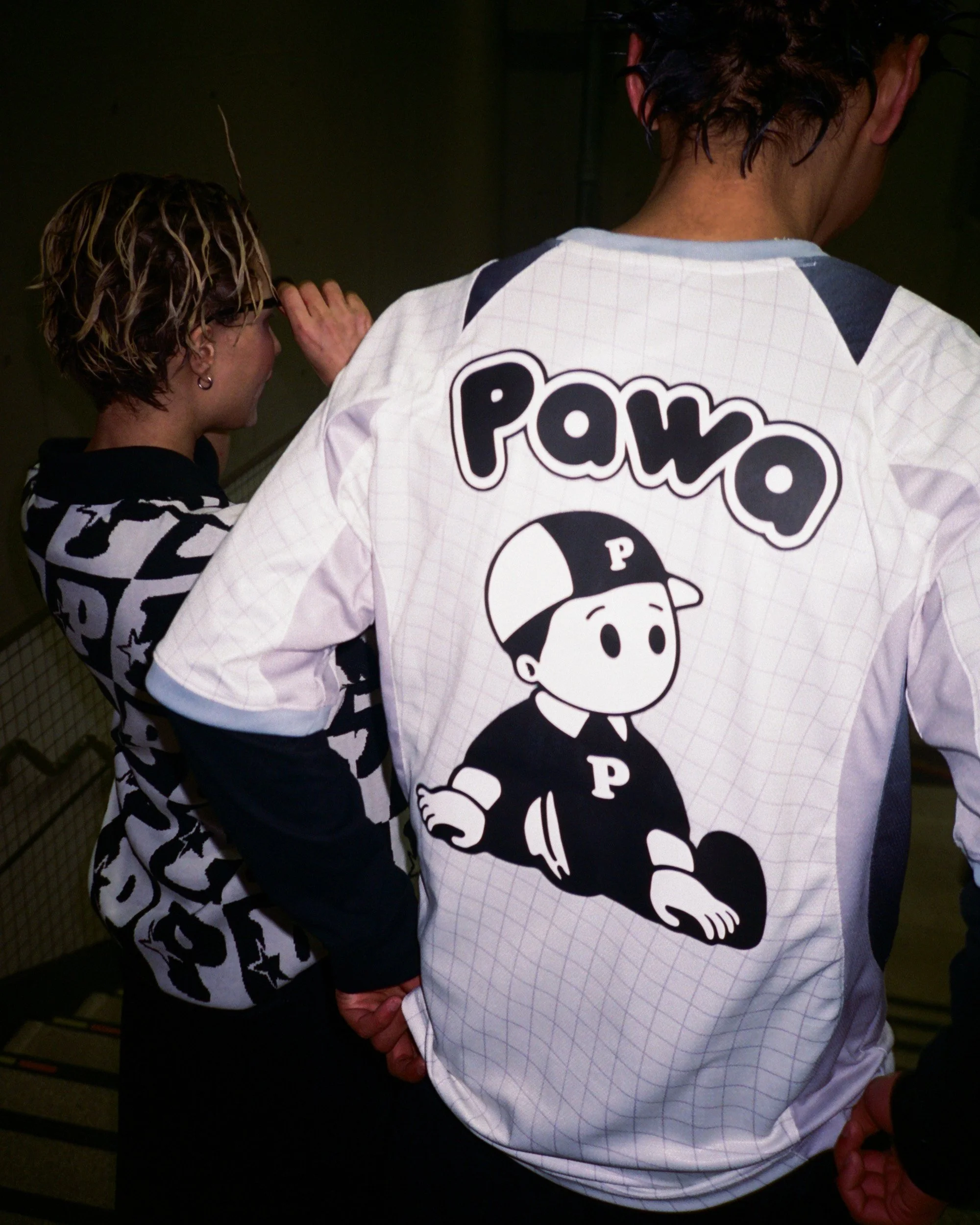 Two people standing close together, one with short hair and the other with longer, curly hair. The person with longer hair has their hand on the other's face. Both are dressed in black and white clothing, with the person facing the camera wearing a s