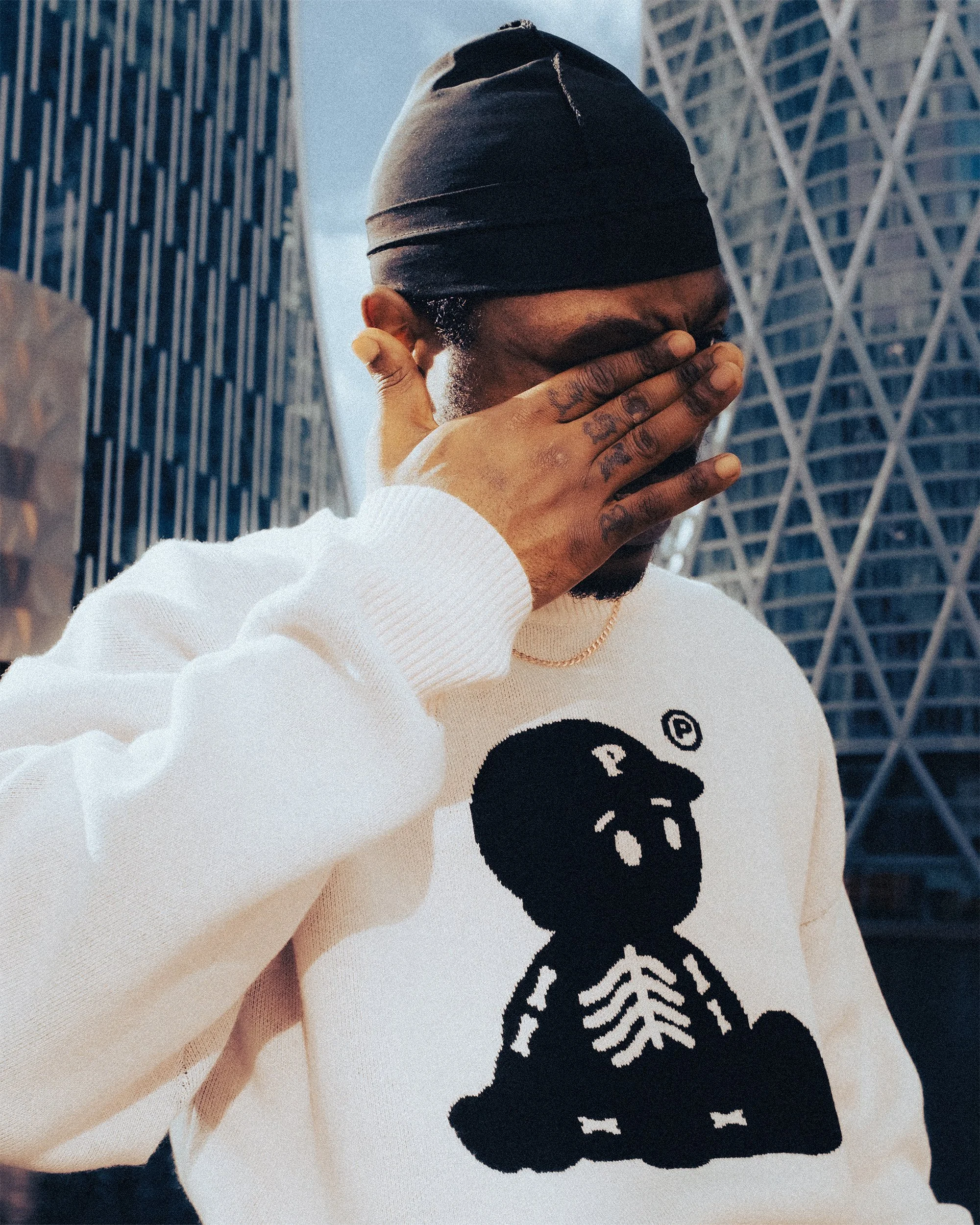 A person wearing a black headscarf and a white sweatshirt with a skull and dollar sign graphic, covering their face with their hand, standing in front of tall buildings.