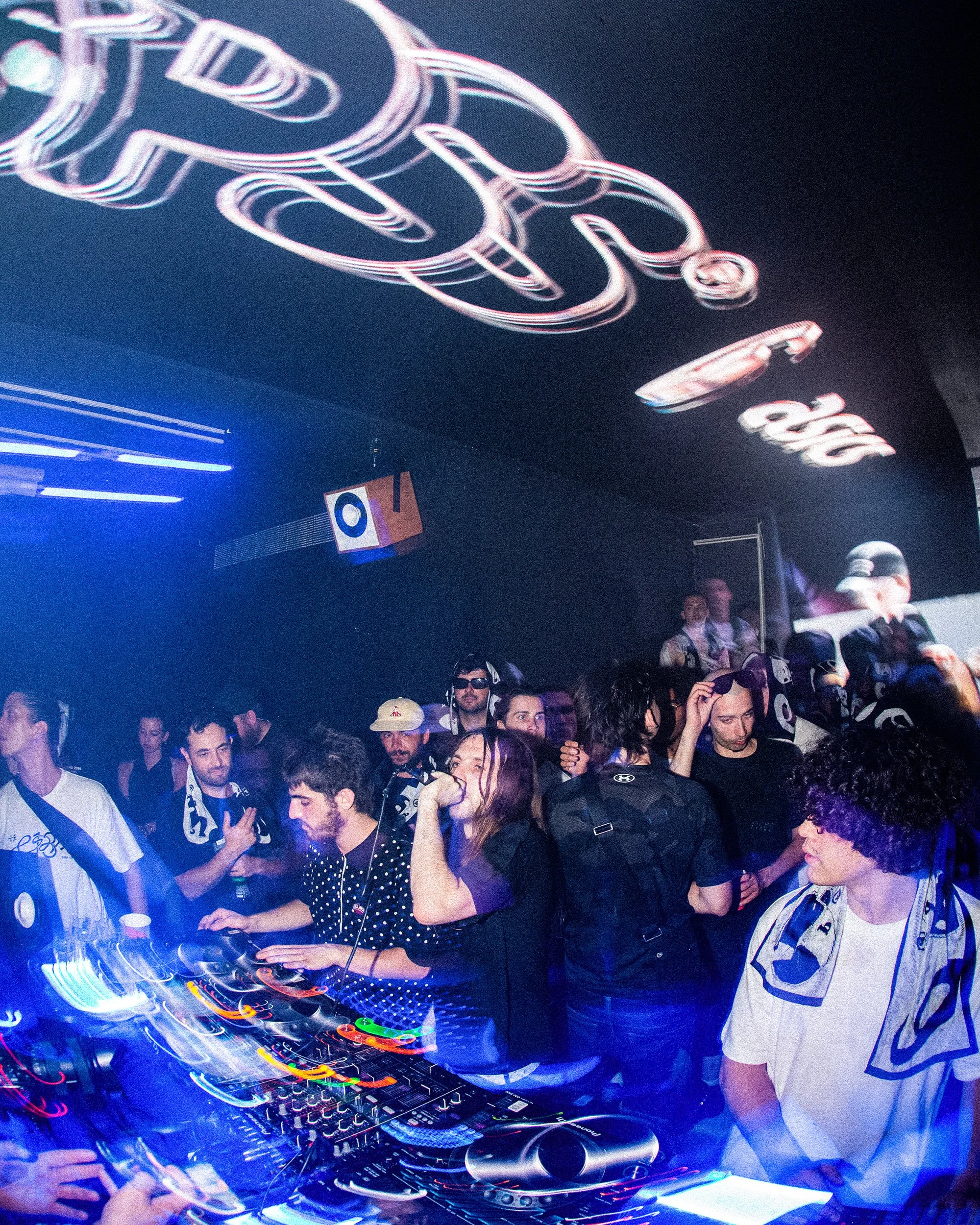 A crowded dance party with people dancing and socializing. There is a DJ setup with colorful lights at the center, and neon signs hanging above. Some attendees are wearing headphones, and others are taking photos or talking.