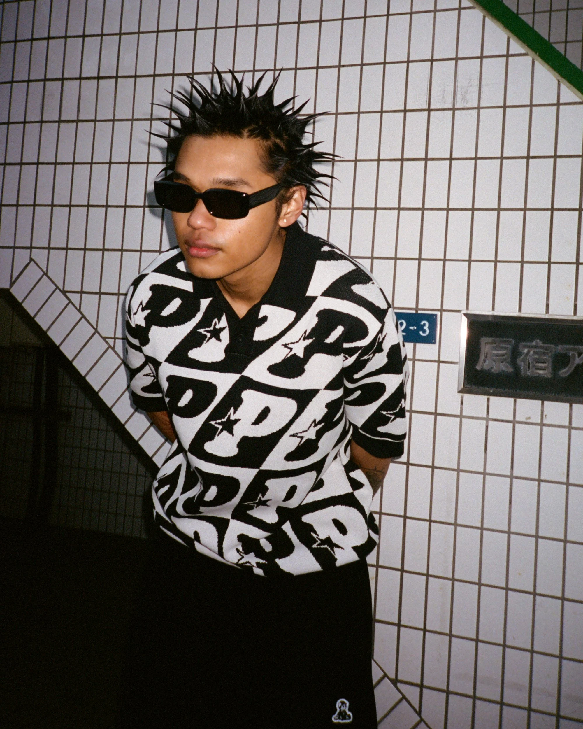 A person with spiky black hair and sunglasses is sitting in front of a white tiled wall with black grid lines, wearing a black and white patterned shirt.