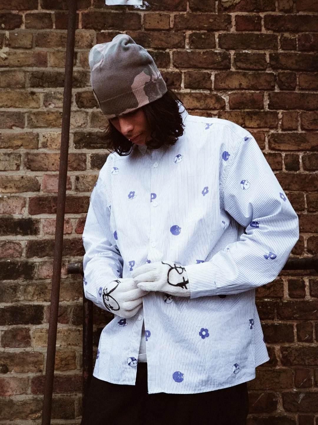 Person wearing a gray beanie, white gloves, and a white shirt with blue flower and letter prints standing against a brick wall.