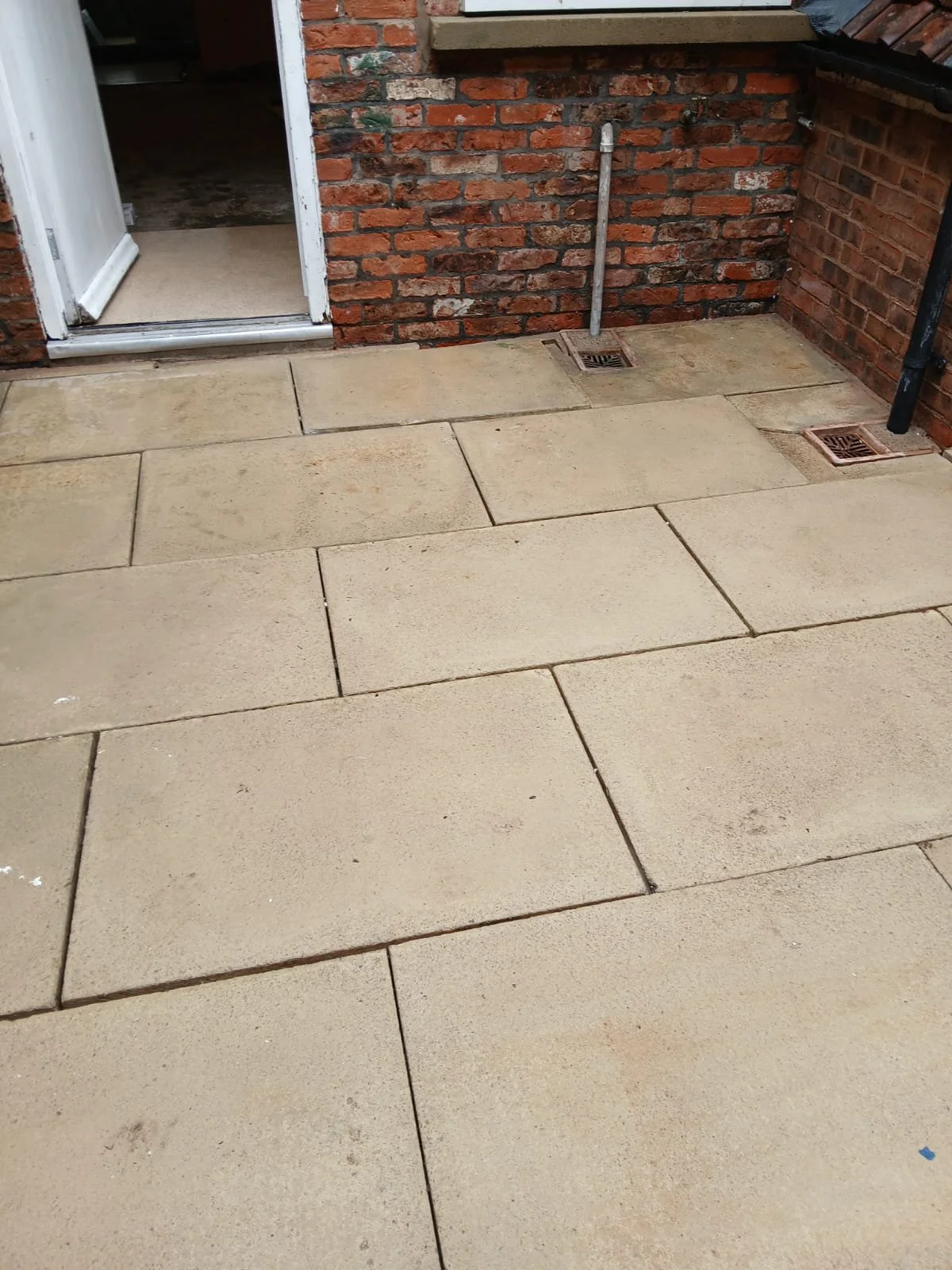Exterior view of a patio with large beige paving stones, brick walls, a white door, a metal pipe, and drain covers.