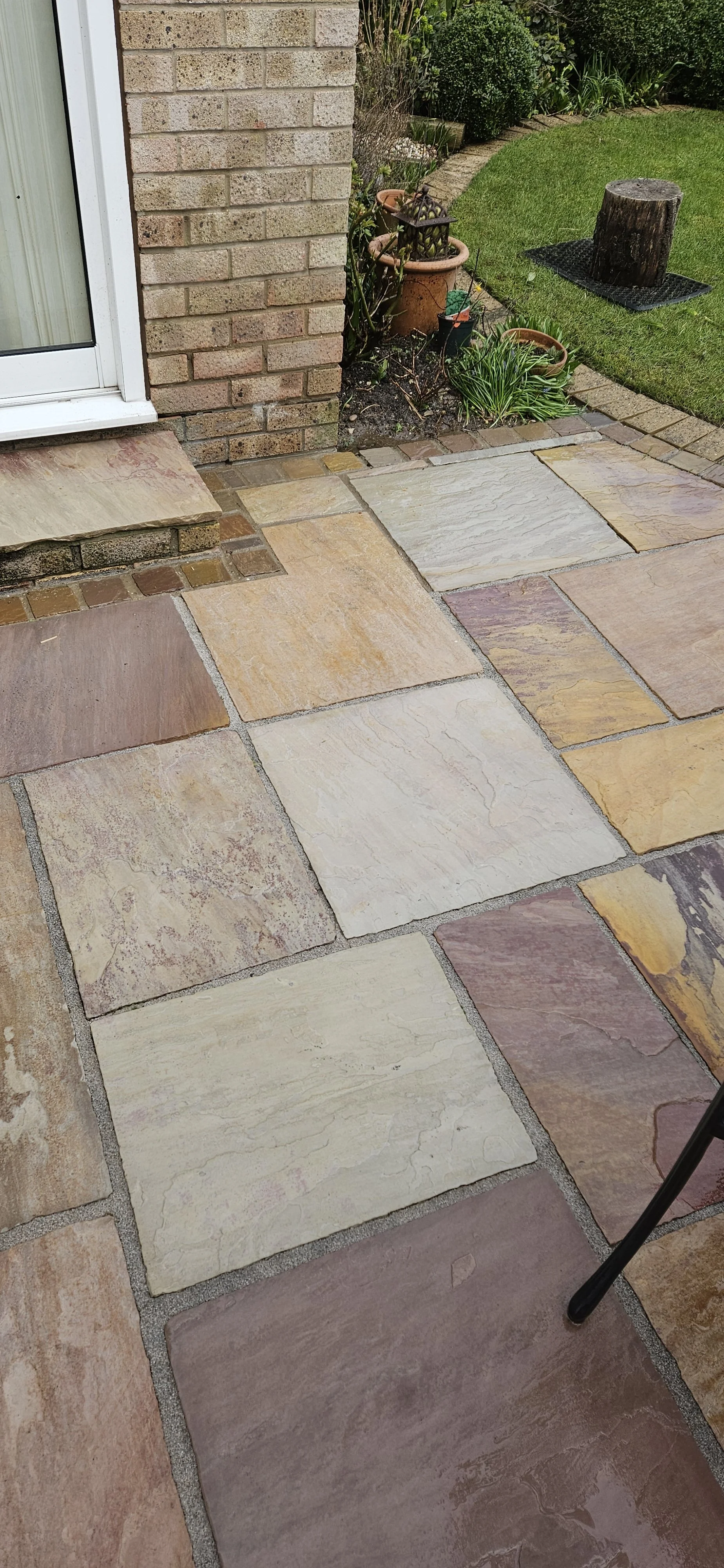 Stone patio with various shades of natural stone slabs; part of a house with brick wall and glass door; small garden with potted plants, a tree stump, and greenery in the background.