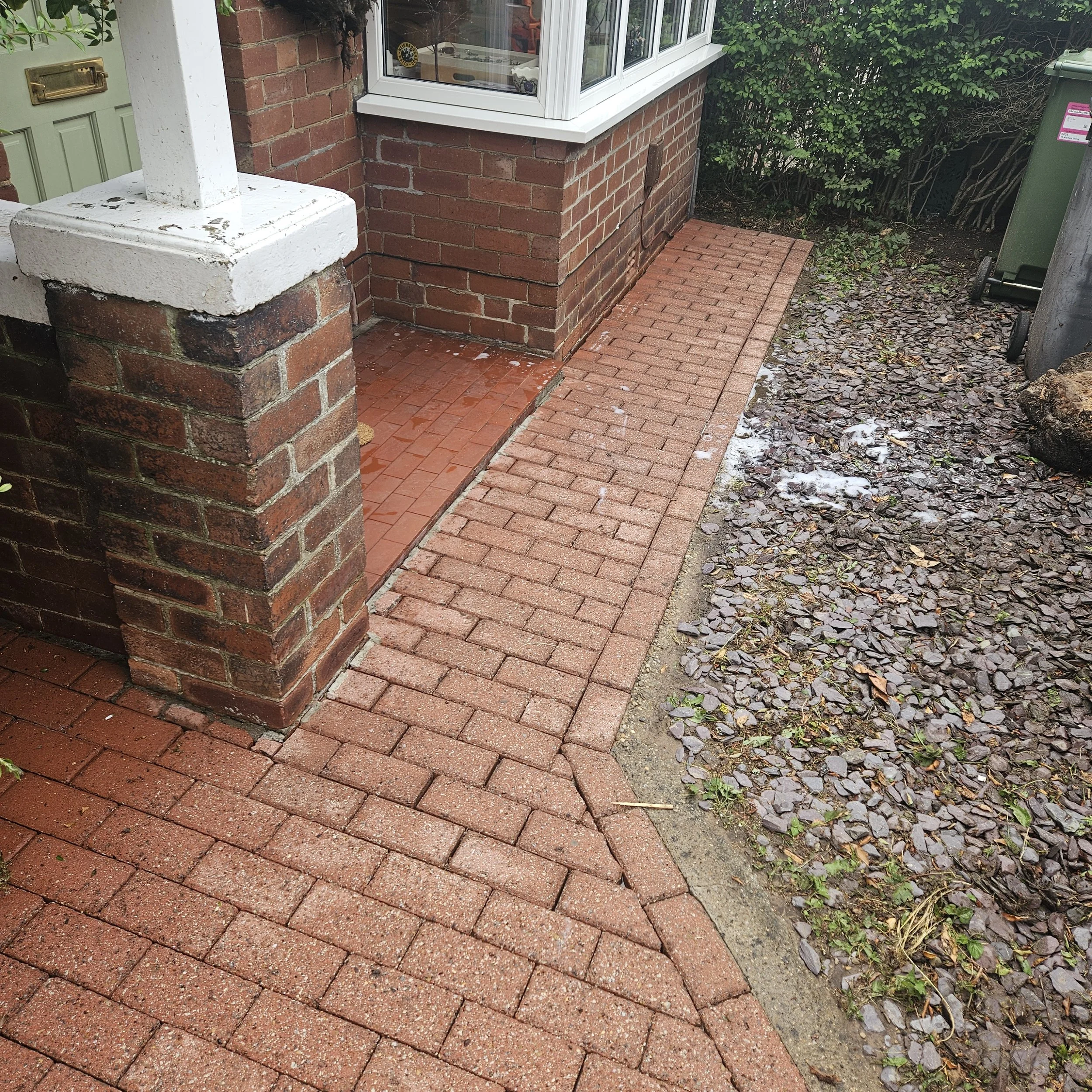 A small front yard with a brick walkway and a brick porch. There is a brick wall at the corner of the house, with a white windowsill and trim. A green gate with a mailbox is visible on the left, and a green garbage bin is on the right. The yard has g