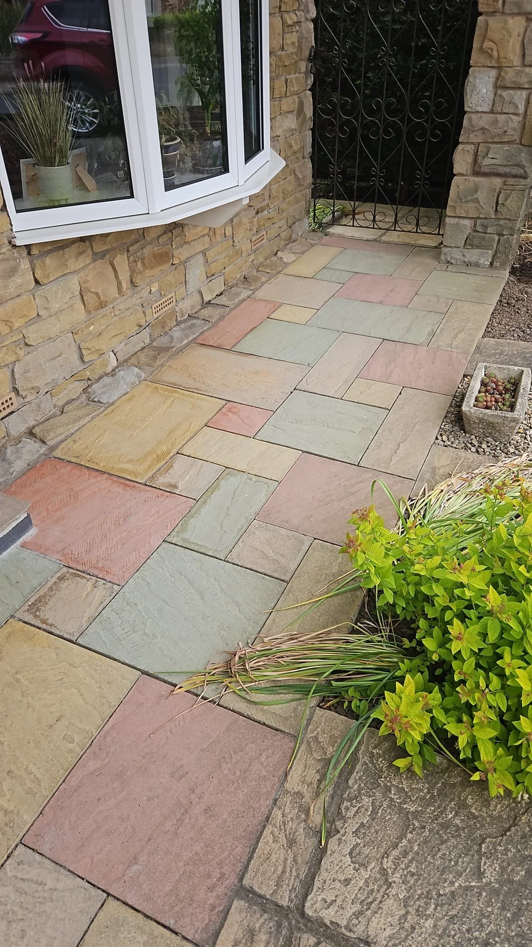 Colorful multicolored stone patio outside a house, with a window and a decorative black metal gate, green plants in a pot and a rectangular concrete planter with succulents.