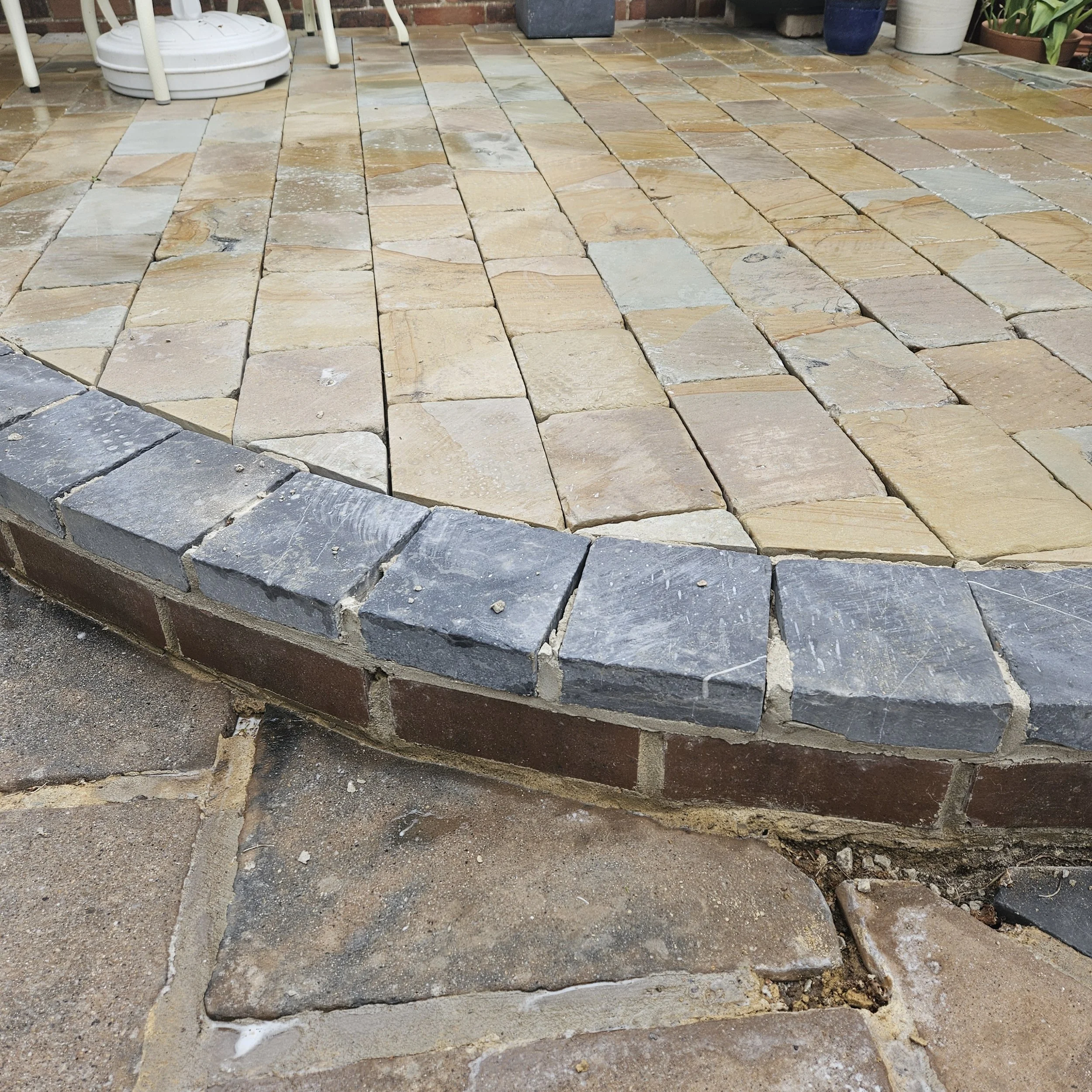 Close-up of a partially installed stone patio with rectangular beige and gray stones, some with visible mortar and gaps, and a construction edge at the bottom.