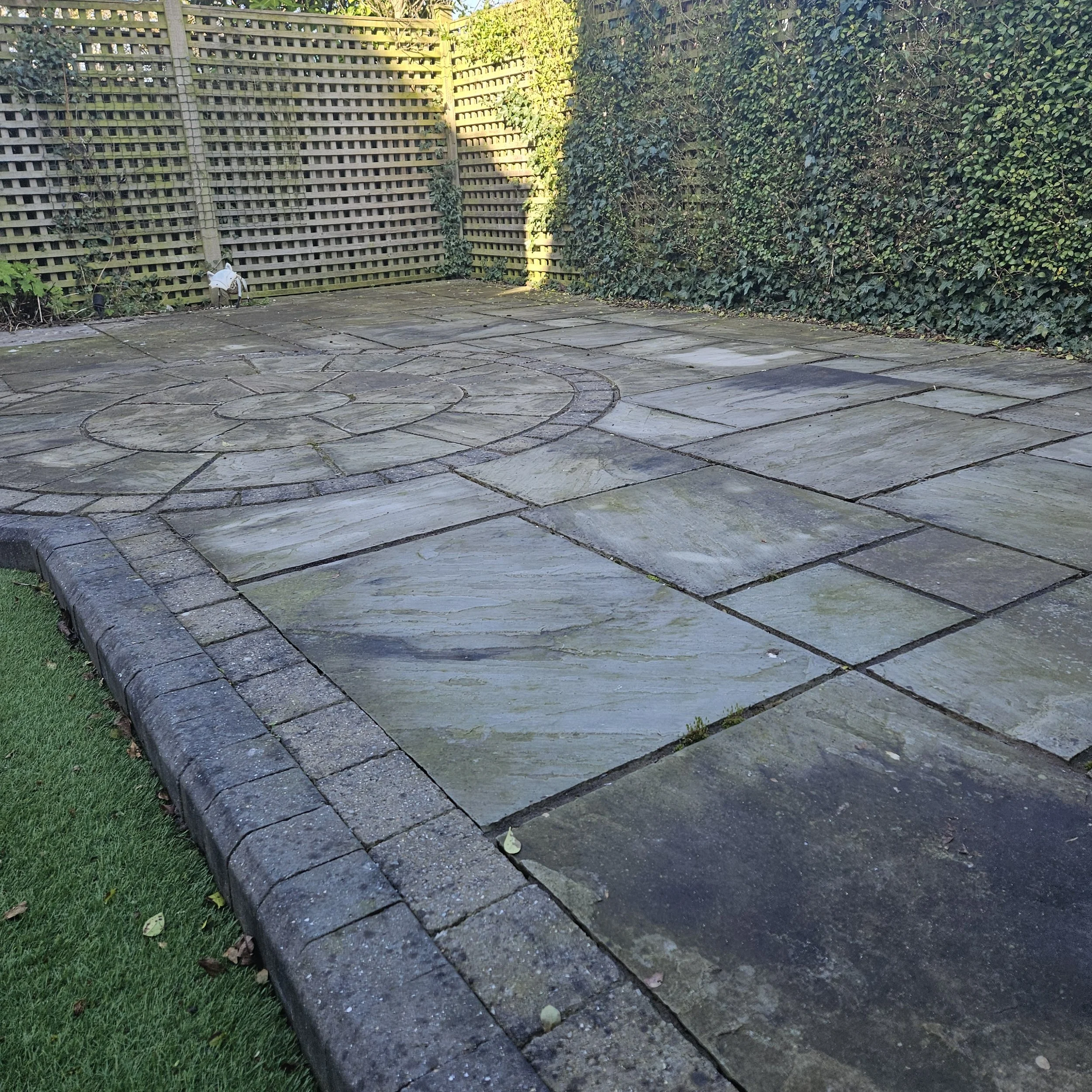 A backyard patio with gray stone pavers bordered by a curved stone edge, a lattice fence on the left, and dense green bushes on the right, with a small garden bed on the left corner and a few scattered leaves on the ground.