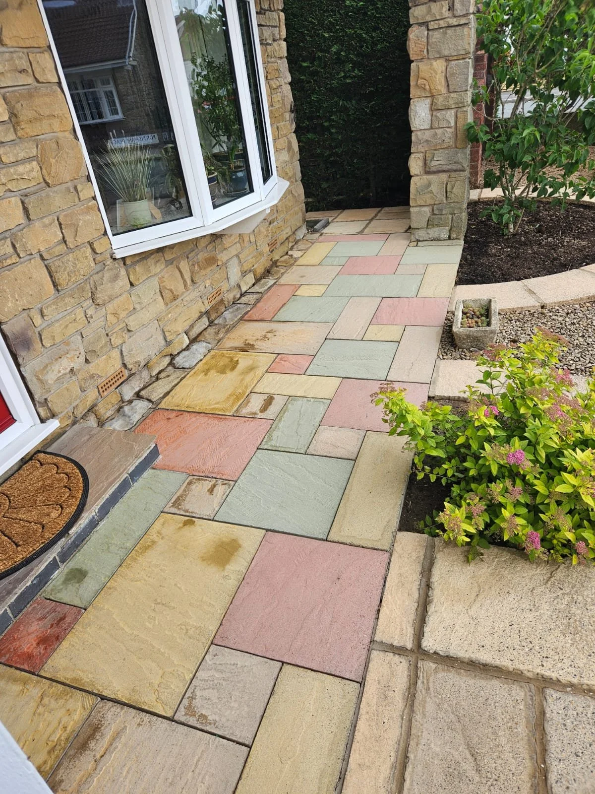 Colorful stone patio leading up to a house with brick exterior walls and a window with potted plants. There are garden beds with green plants and flowers along the edge.