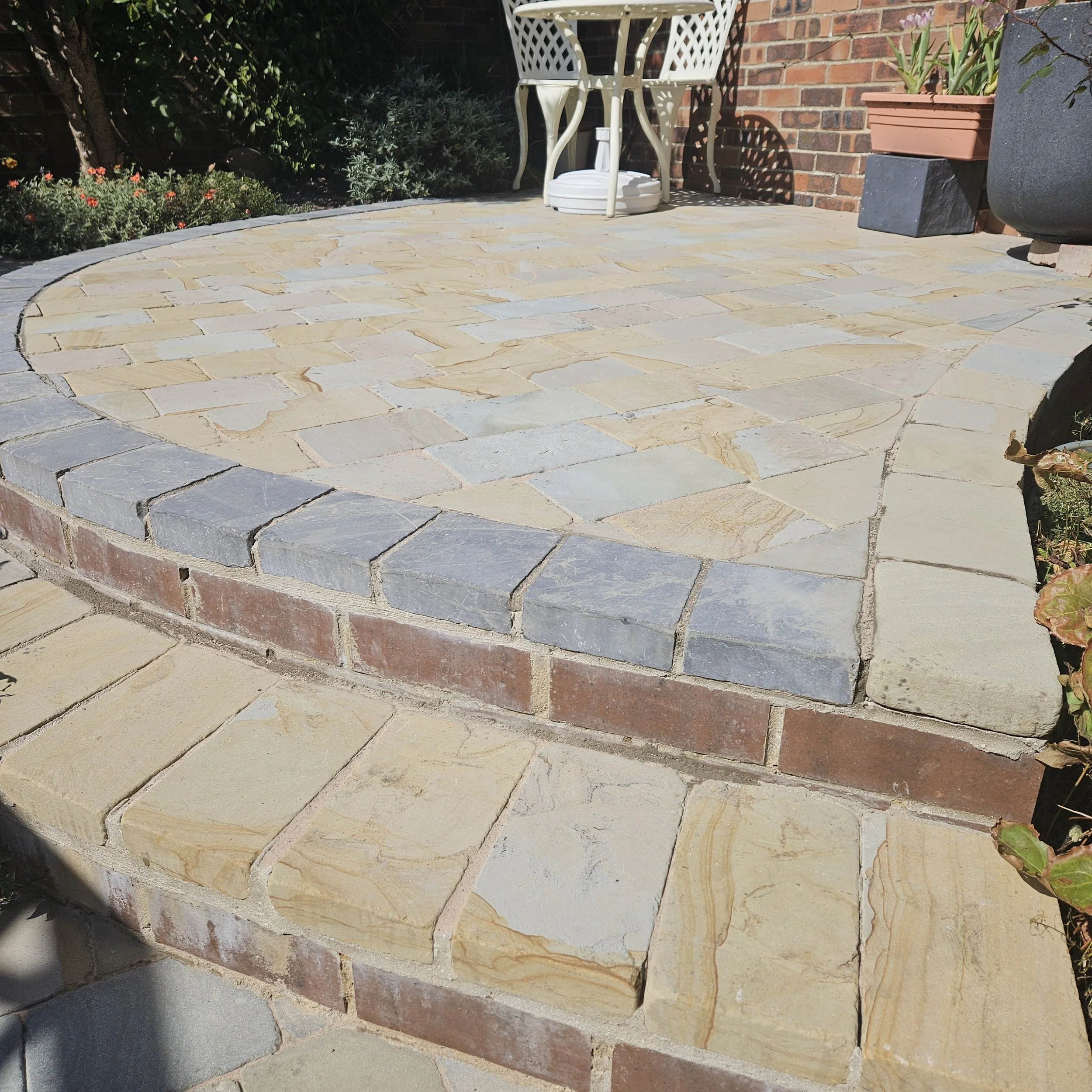 A curved section of a stone patio with multicolored rectangular stones in shades of yellow, beige, pink, and blue. There is a brick wall with potted plants and garden greenery in the background.
