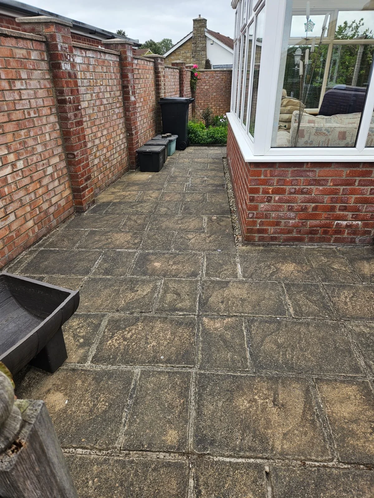 Backyard patio with weathered stone tiles, brick wall on left, brick and glass conservatory on right, and a small garden with pink flowers and green plants in the background.