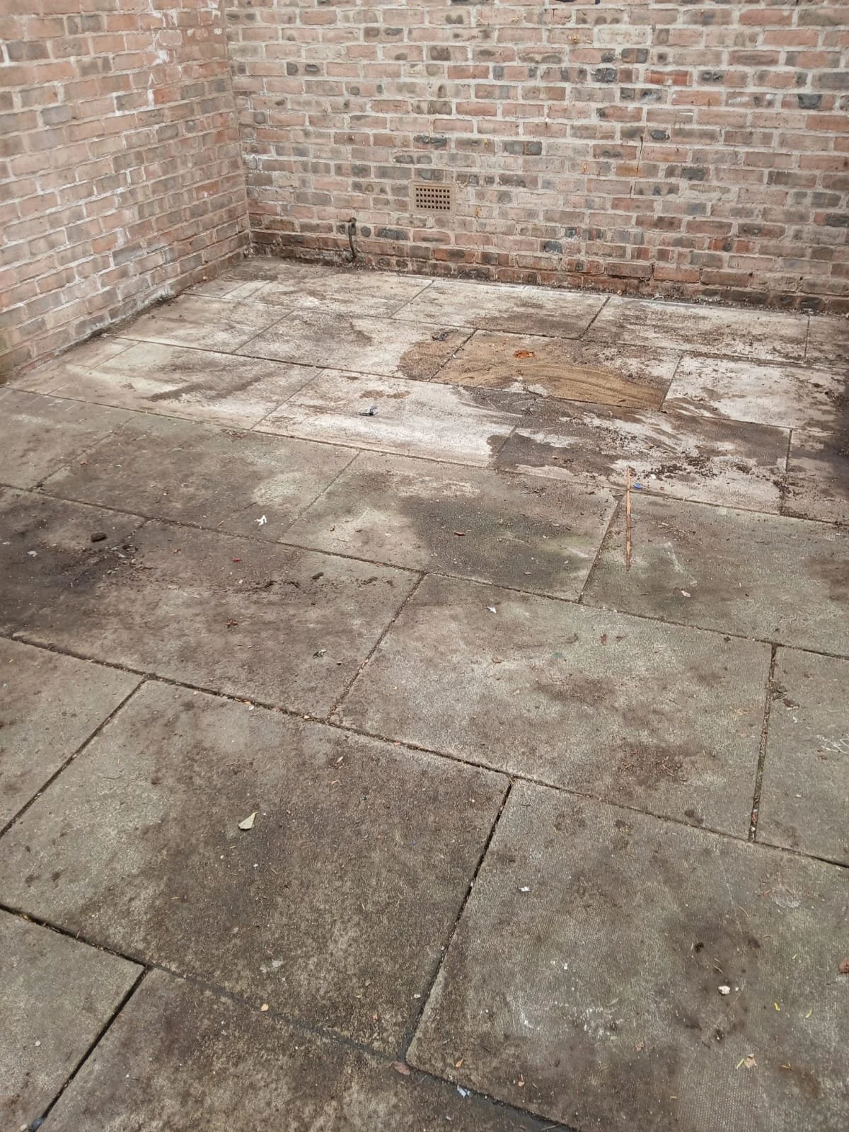 The image shows an outdoor concrete patio area with large, weathered concrete slabs. There are brick walls with some pipes and a vent on one side, and the ground has dirt, small debris, and signs of wear.