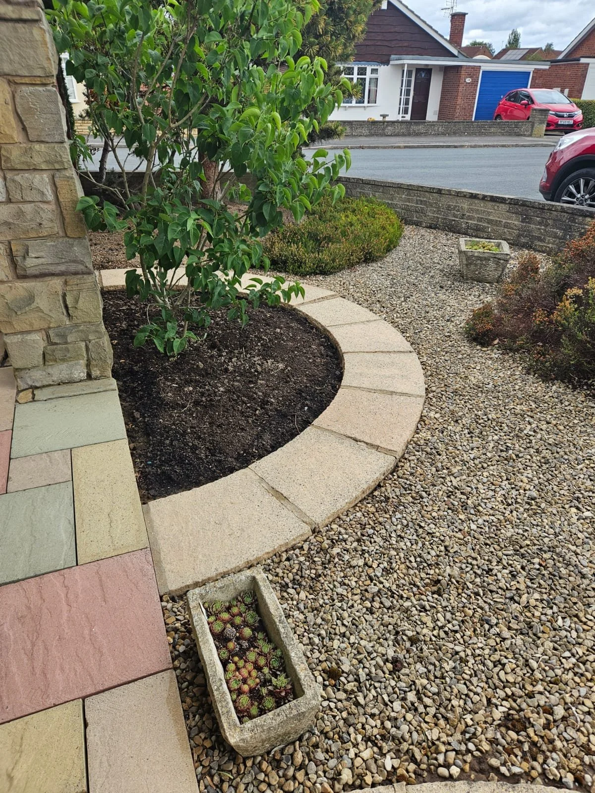 A landscaped garden with a curved stone border, bushes, and a small tree, with a pebble pathway and a row of houses in the background.
