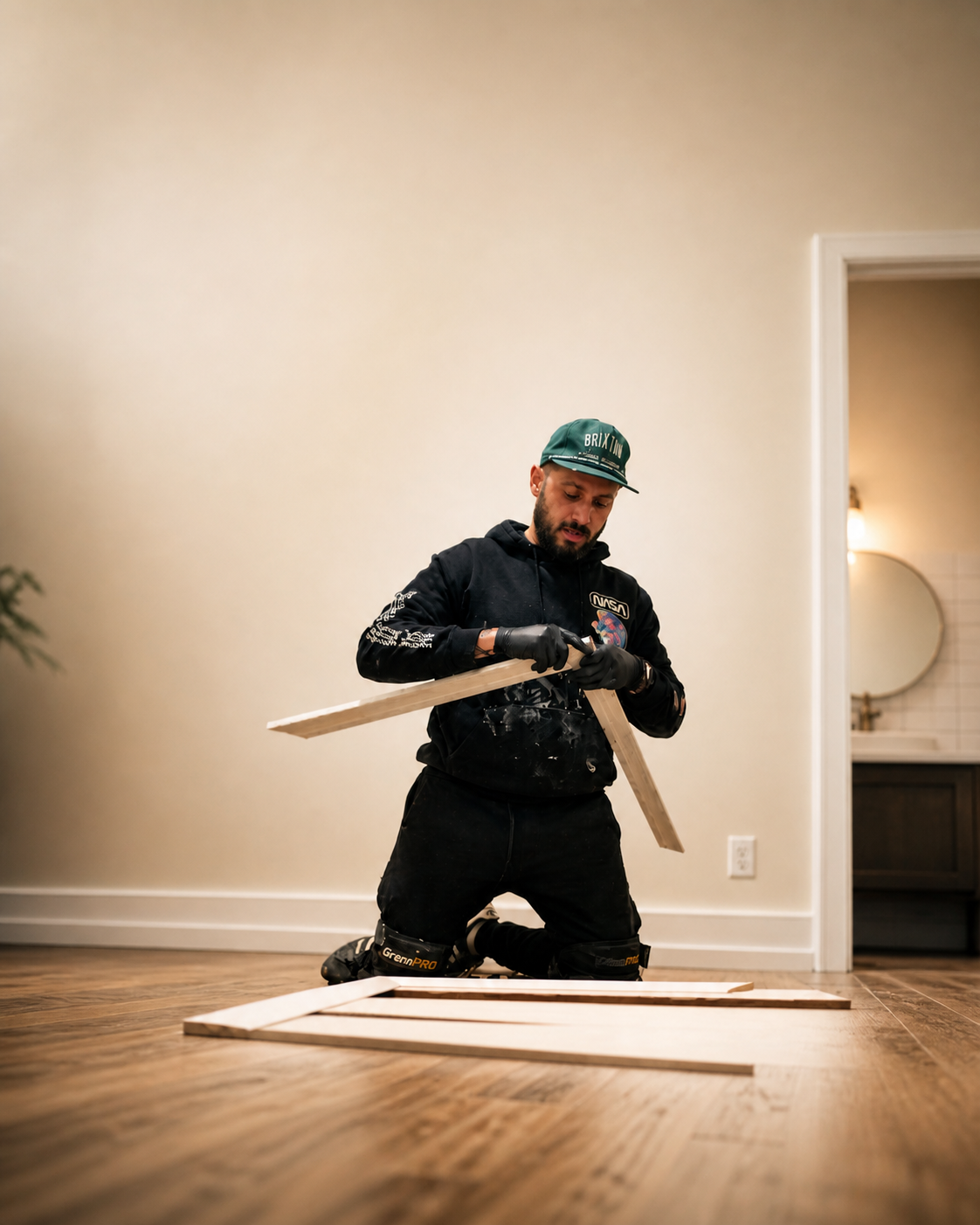 A man kneeling on the wooden floor, assembling or working on wooden frame pieces. He is wearing a black hoodie, black gloves, a green cap, and kneepads, in a room with a white wall, a doorway leading to a bathroom with a sink and mirror.