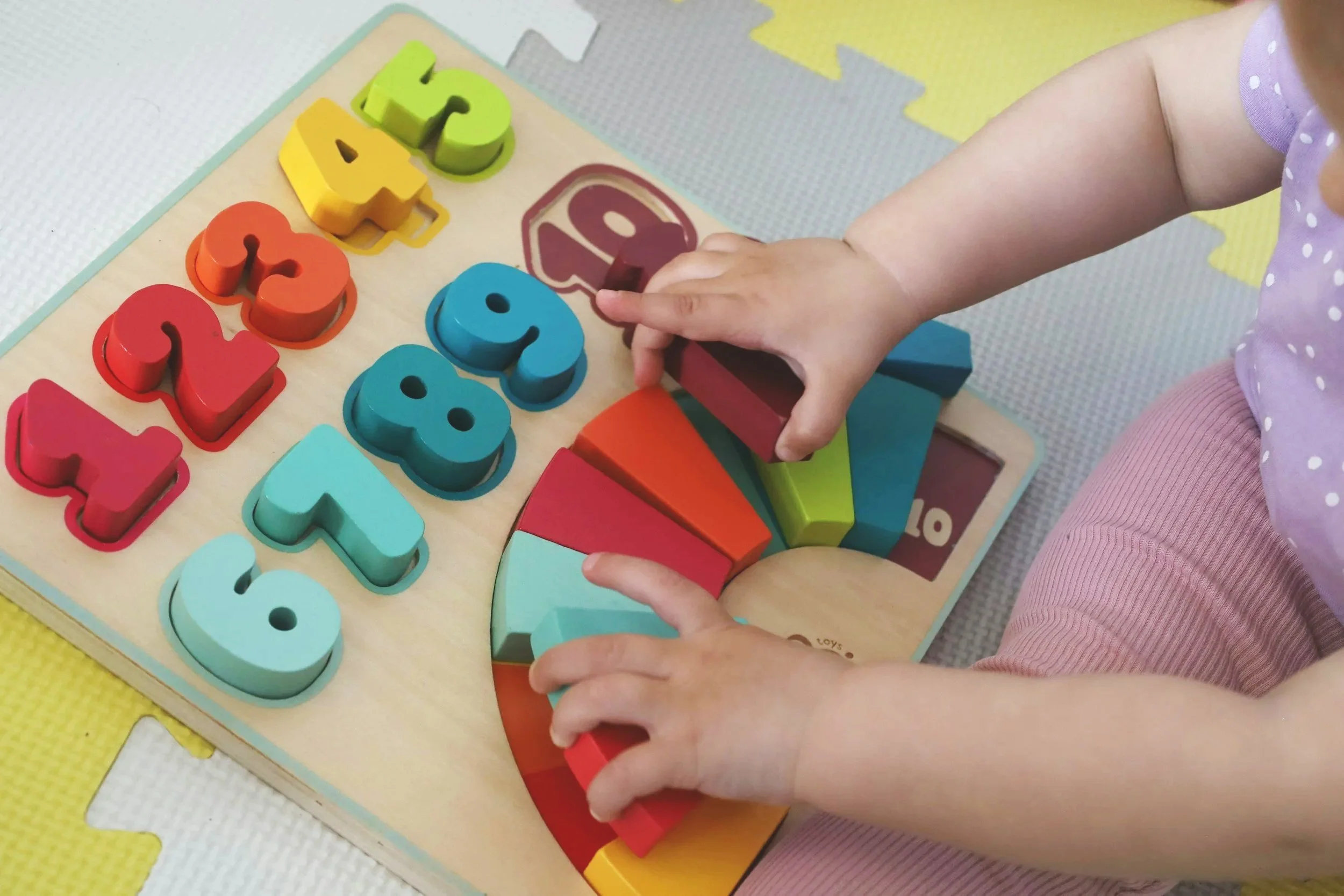 Child's hands placing wooden number puzzle pieces on a board, with colorful numerals and shapes.