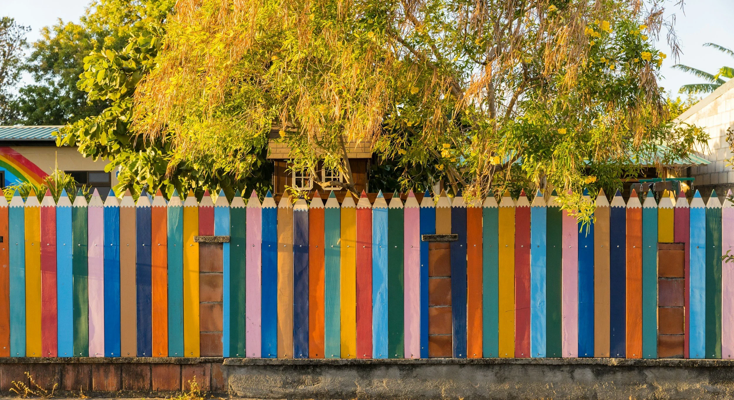 Colorful wooden fence in front of a house with a large leafy tree and a part of a rainbow mural on the wall.