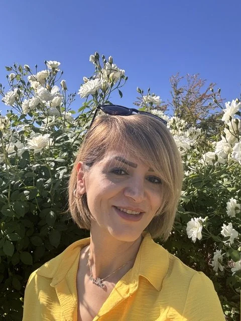 Woman with short blonde hair smiling outdoors in front of white flowering bushes, wearing a yellow jacket and sunglasses on her head.
