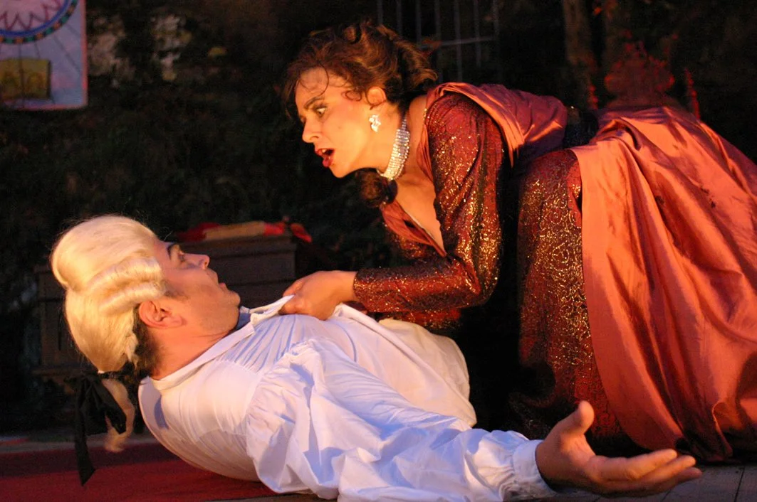 Two women, one lying on the ground and the other leaning over her, engaged in a heated conversation or argument on a stage with a dark outdoor background.