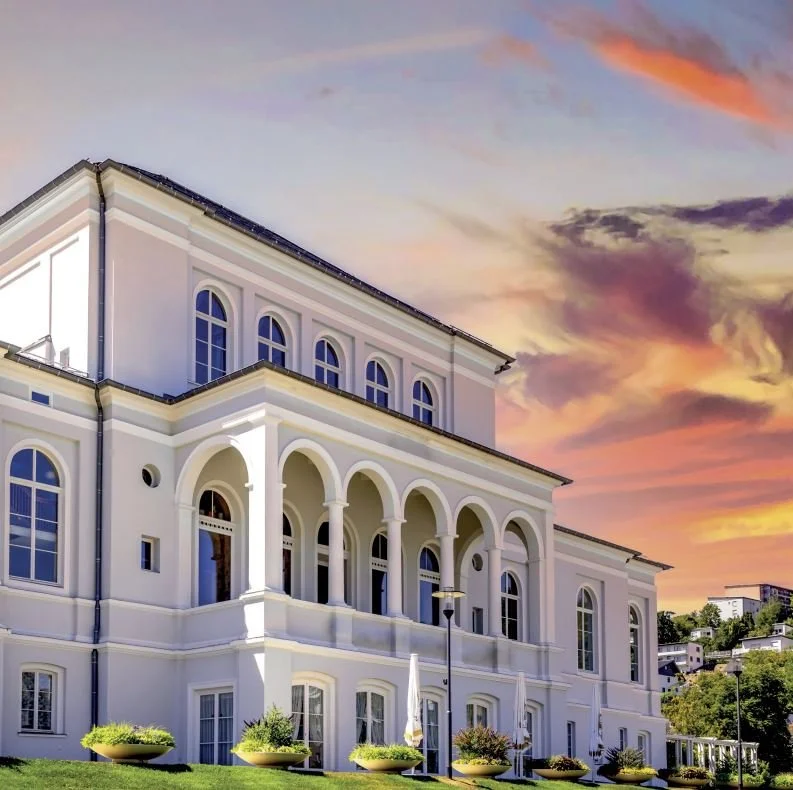 A large white neoclassical building with arched windows and columns at sunset, with a colorful sky and landscaped garden in front.