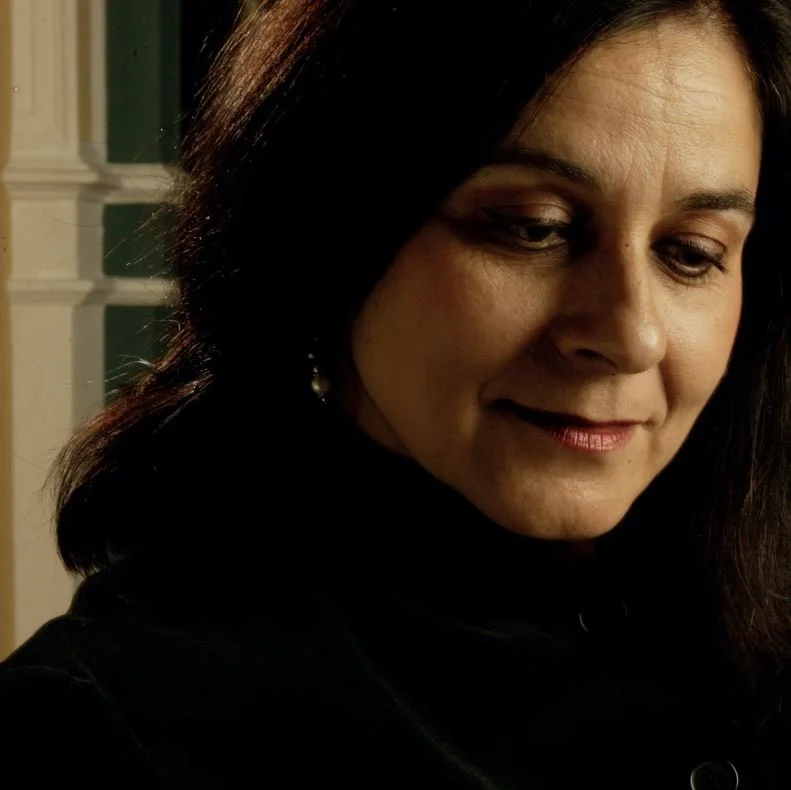 Close-up of a woman with dark hair, looking down and smiling softly, in a dimly lit room with a green and white background.