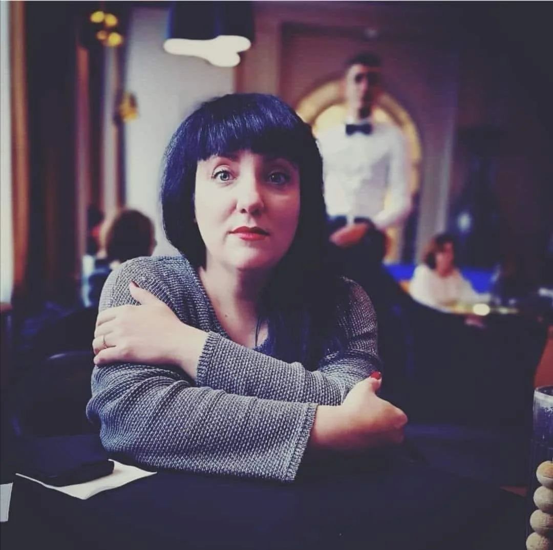 A woman with shoulder-length dark hair and light skin sitting at a table in a restaurant, with her arms crossed and looking directly at the camera.