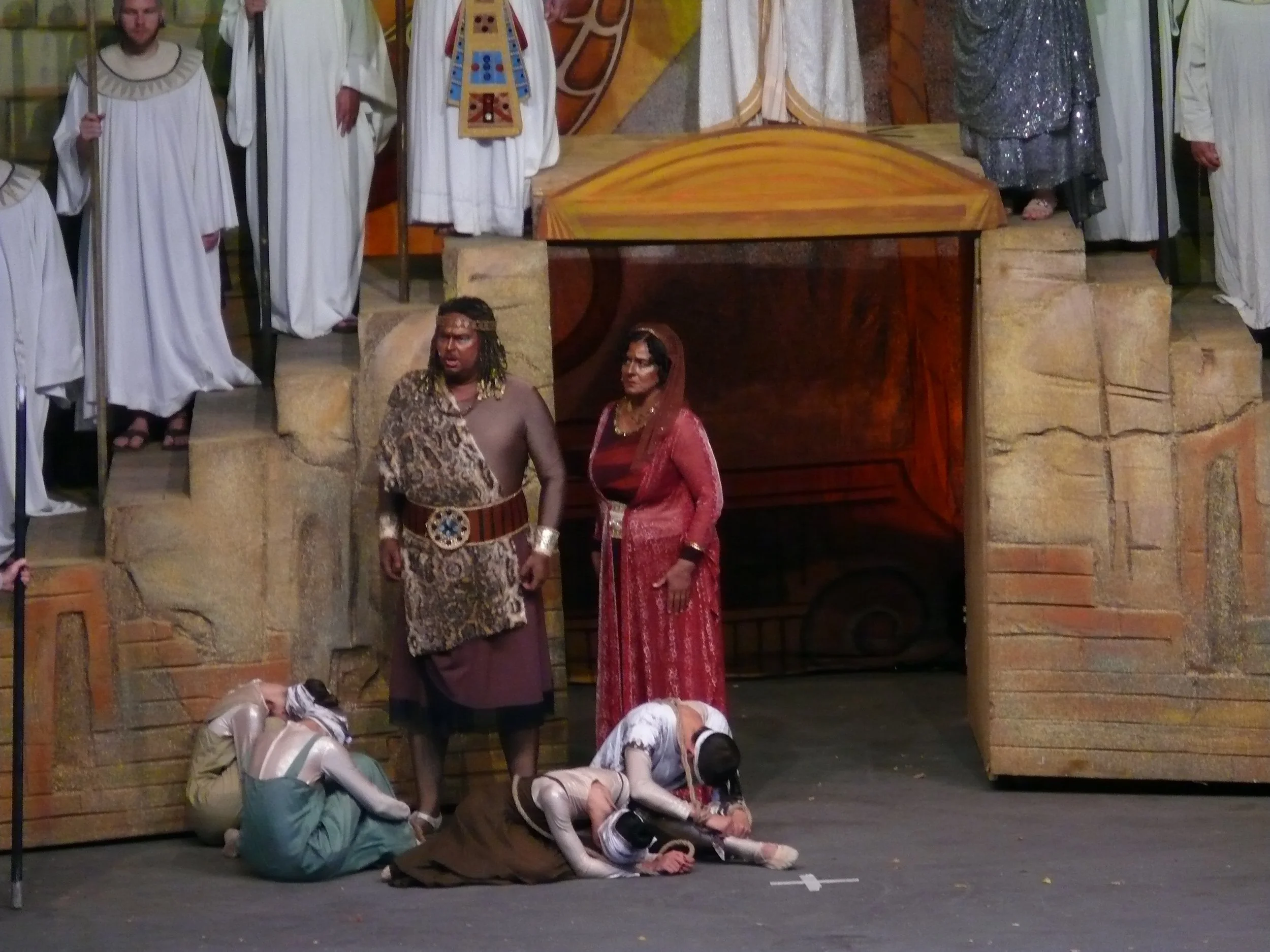 People dressed in costumes performing a dramatic scene on a wooden stage with stone props and a painted backdrop, depicting a scene from a theatrical play.