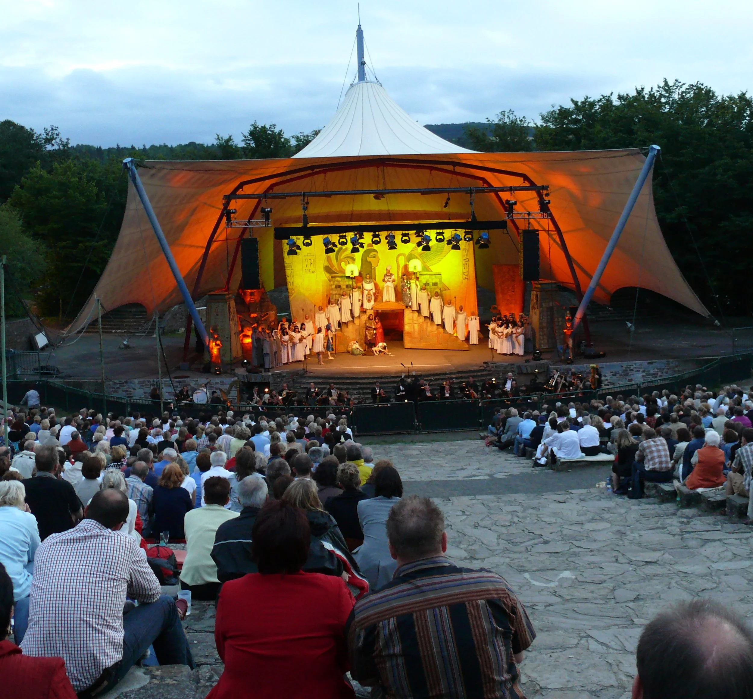 Outdoor auditorium or stage with tents and people gathered for a performance or event at sunset.