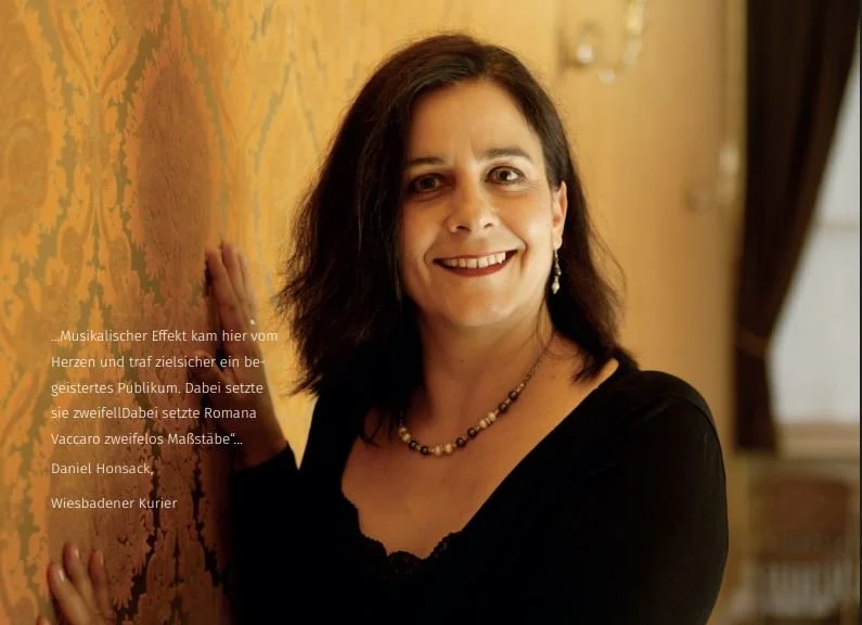 A woman with dark hair smiling and posing in a black top, standing next to a yellow patterned wall in a room with curtains.