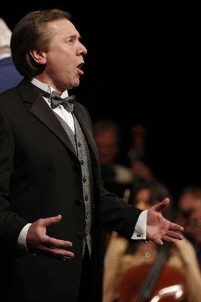 A male opera singer performing on stage, dressed in formal black tuxedo with a bow tie, with an audience in the background.