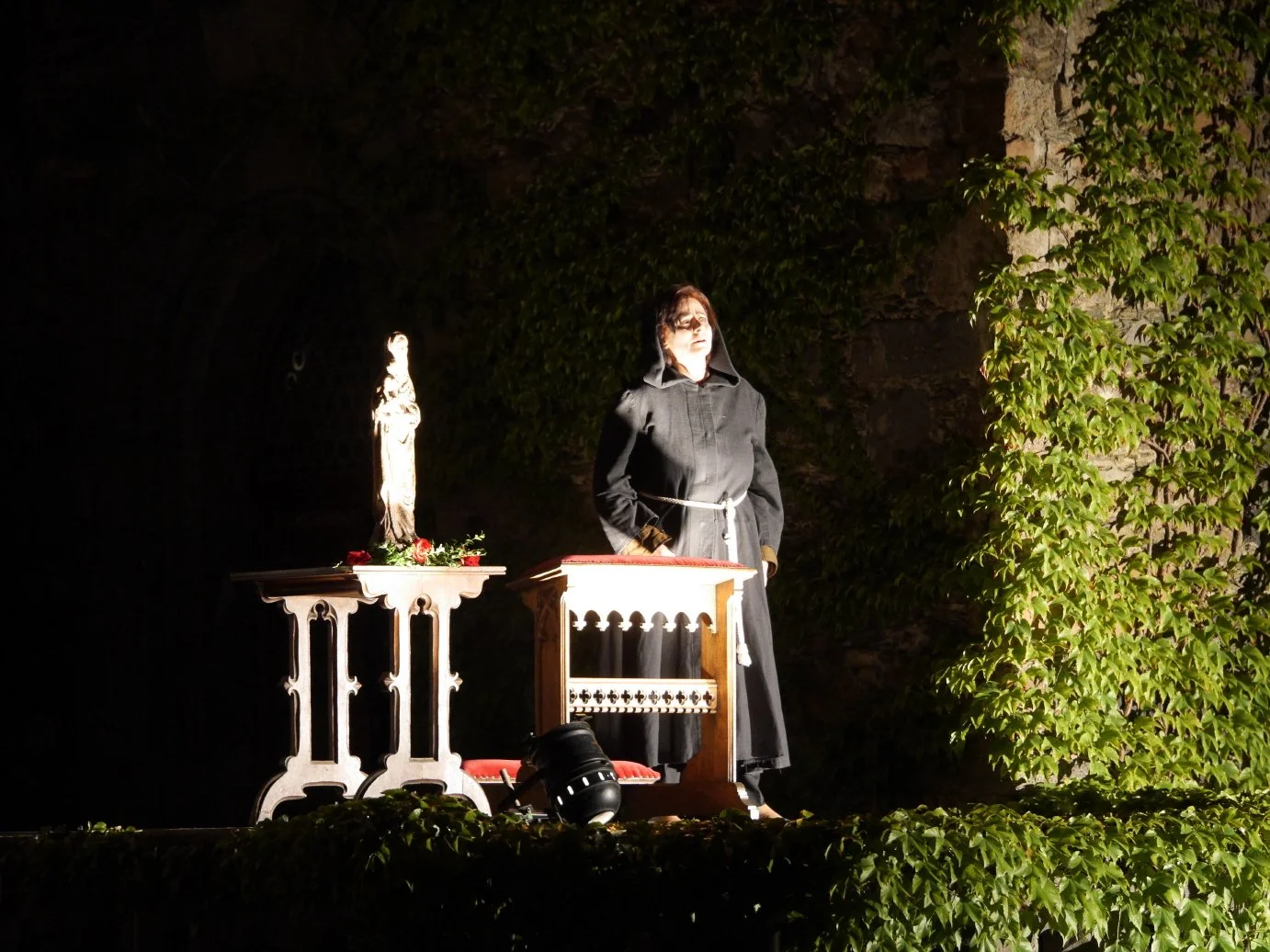 A woman in a black coat standing outdoors at dusk, next to a table with a religious statue and roses, surrounded by greenery and an illuminated stone wall.